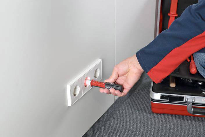 Osoba przeprowadza kontrolę bezpieczeństwa w gniazdku elektrycznym za pomocą testera napięcia. Obok stoi otwarty kufer narzędziowy.