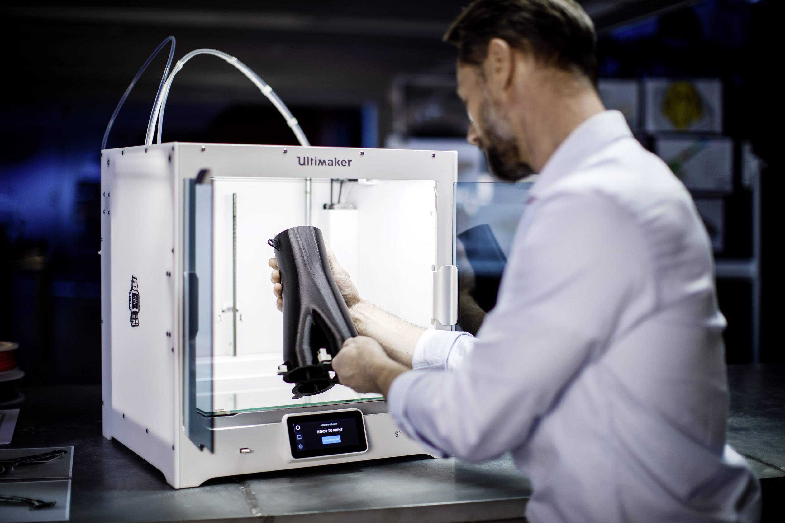 Mężczyzna wyjmuje czarny obiekt z drukarki 3D w laboratorium. Drukarka jest biała i elektronicznie podświetlona.