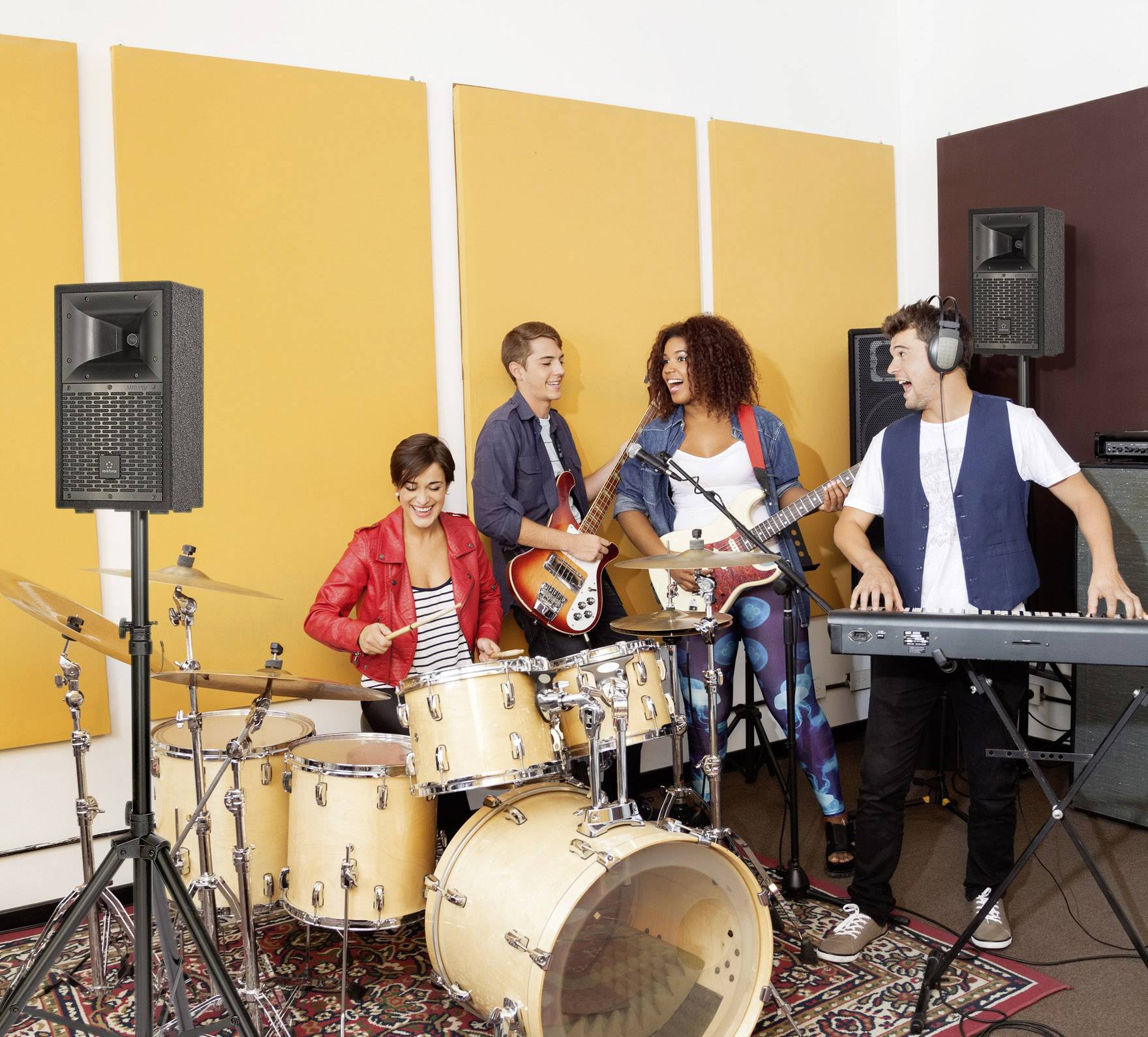 Grupa młodych muzyków ćwiczy w studio. Jedna osoba gra na perkusji, inni grają na gitarze, basie i keyboardzie. Wszyscy są radośni.