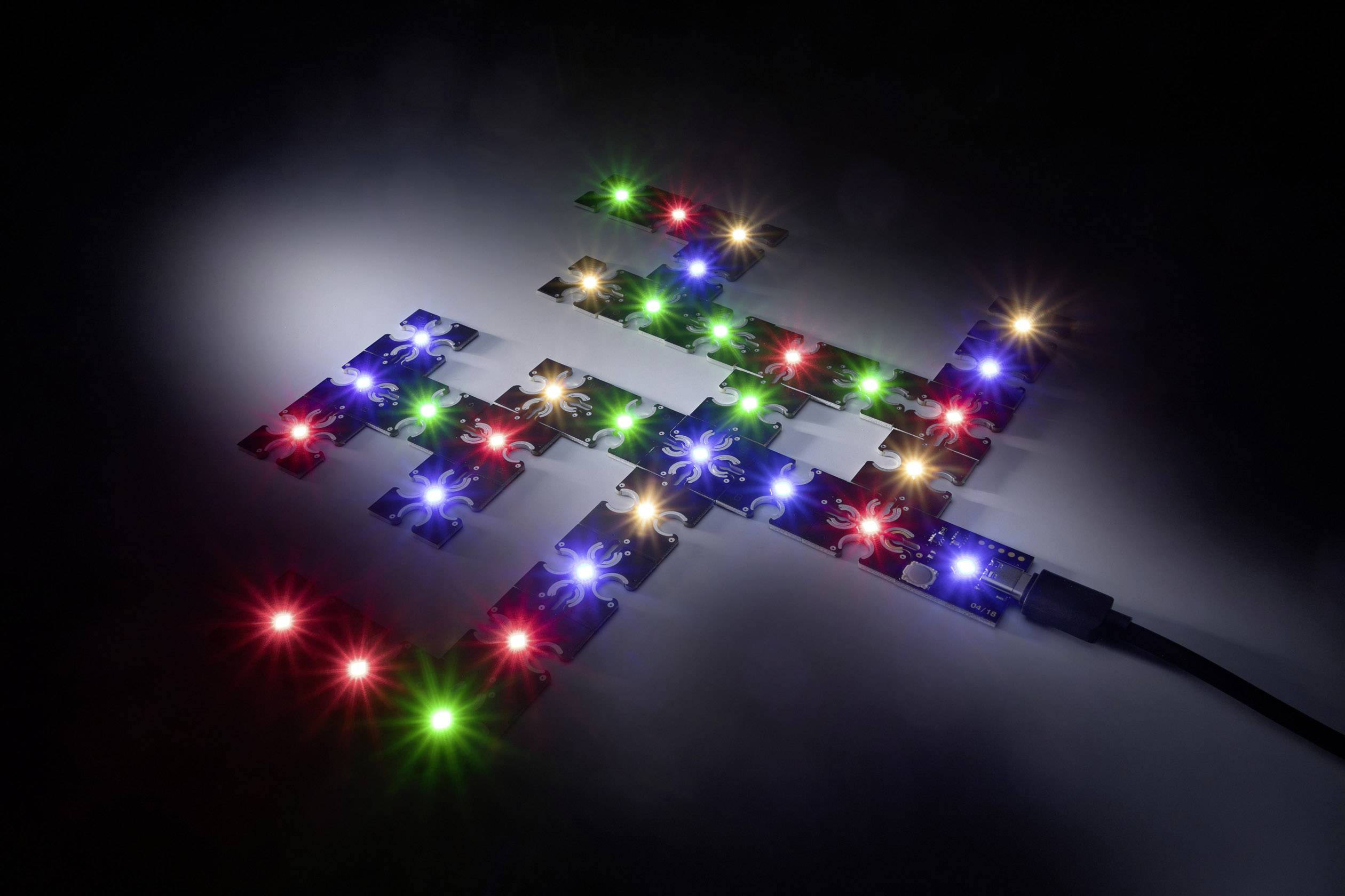 Układ puzzli w kolorze niebieskim, ozdobiony kolorowymi diodami LED w różnych odcieniach, umieszczony na ciemnym tle, tworzący wrażenie technicznej kompozycji.