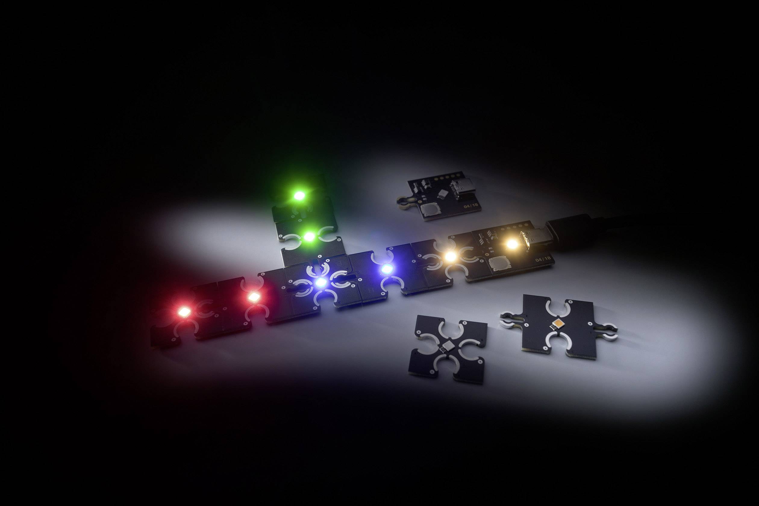 Różne świecące elementy puzzli LED ułożone w ciemności, z czerwonymi, zielonymi, niebieskimi i żółtymi światłami, na czarnym tle.