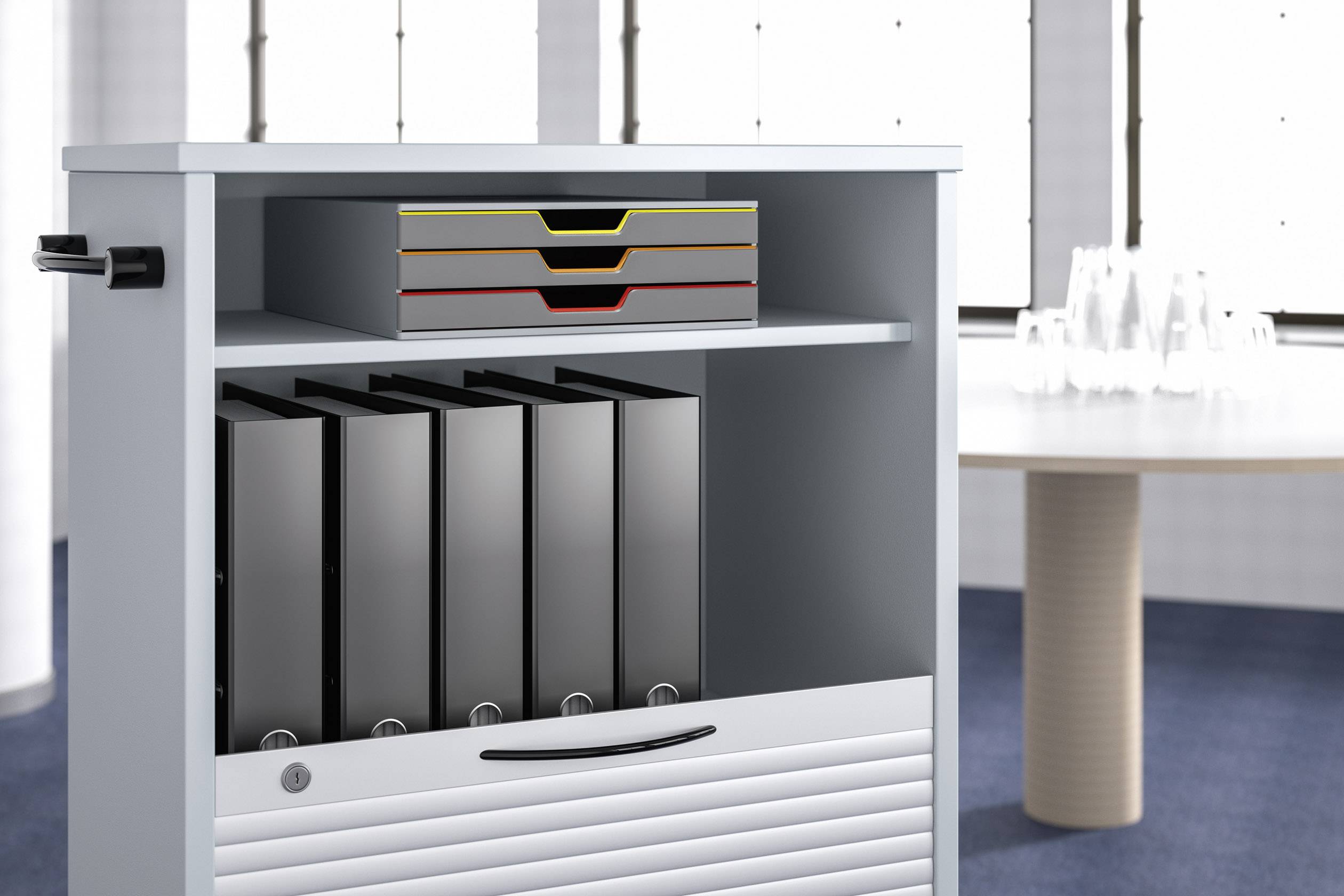 Regał biurowy z pięcioma czarnymi segregatorami i trzema kolorowymi tackami stoi w pomieszczeniu. W tle znajduje się stół z kieliszkami.