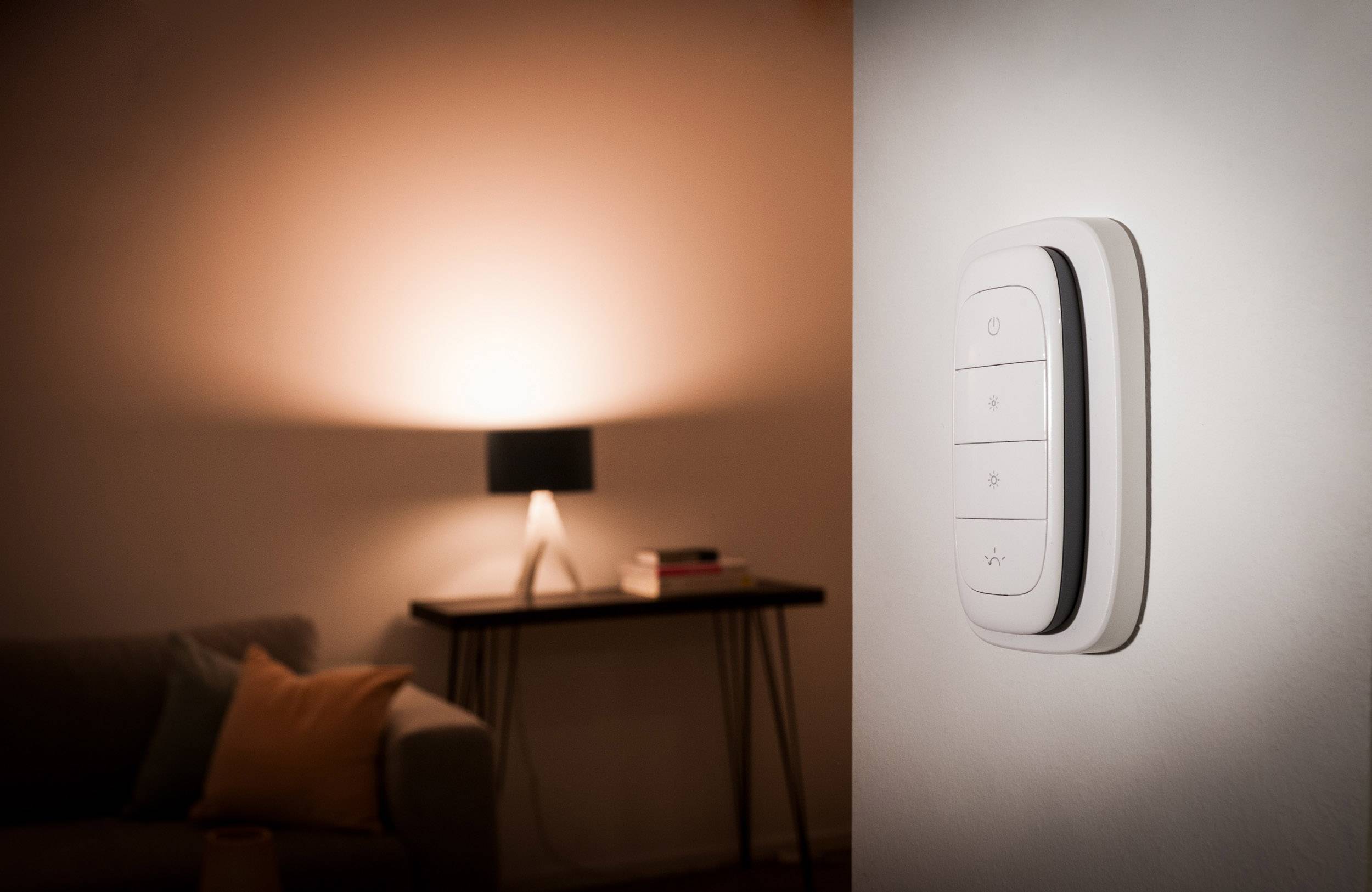 Inteligentny przełącznik świetlny jest zamontowany na ścianie; w tle świeci stylowa lampa stołowa, tworząc przytulną atmosferę.