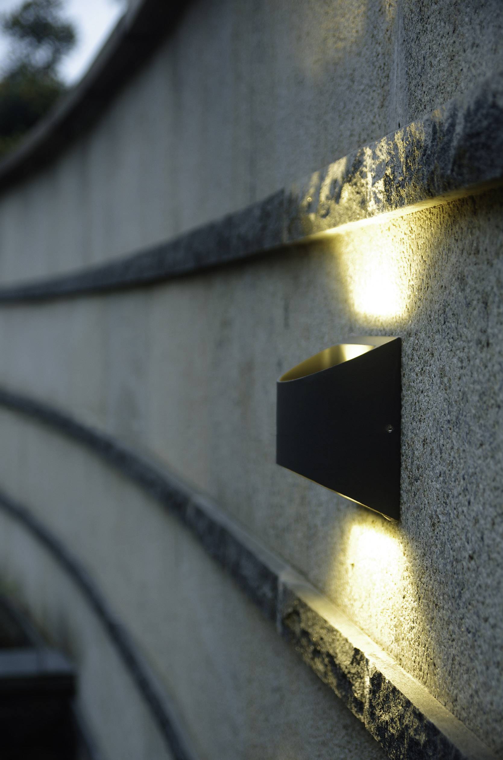 Oświetlona lampa naścienna rzuca ciepłe światło na strukturalny kamienny mur o zmierzchu, tworząc przytulną atmosferę.