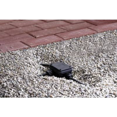 Czarny elektroniczny pudełkowaty sprzęt leży na żwirze obok wybrukowanej ścieżki. Kable biegną po obu stronach urządzenia.
