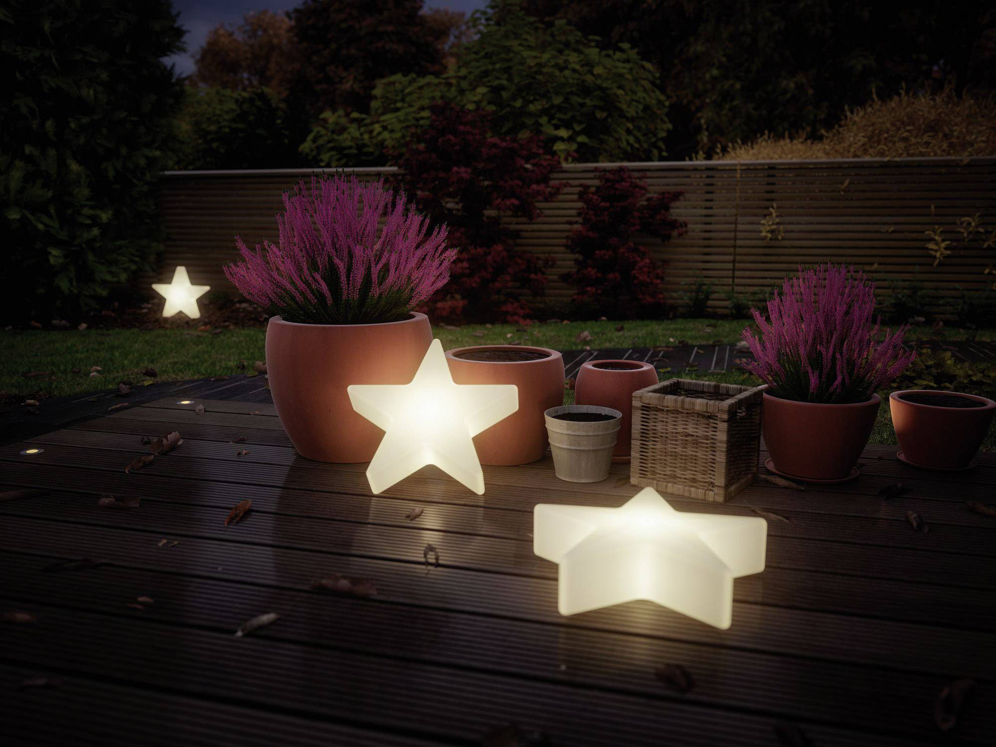Ogród o zmierzchu, z jaśniejącymi lampami w kształcie gwiazd oraz donicami z fioletowymi roślinami na drewnianym tarasie.
