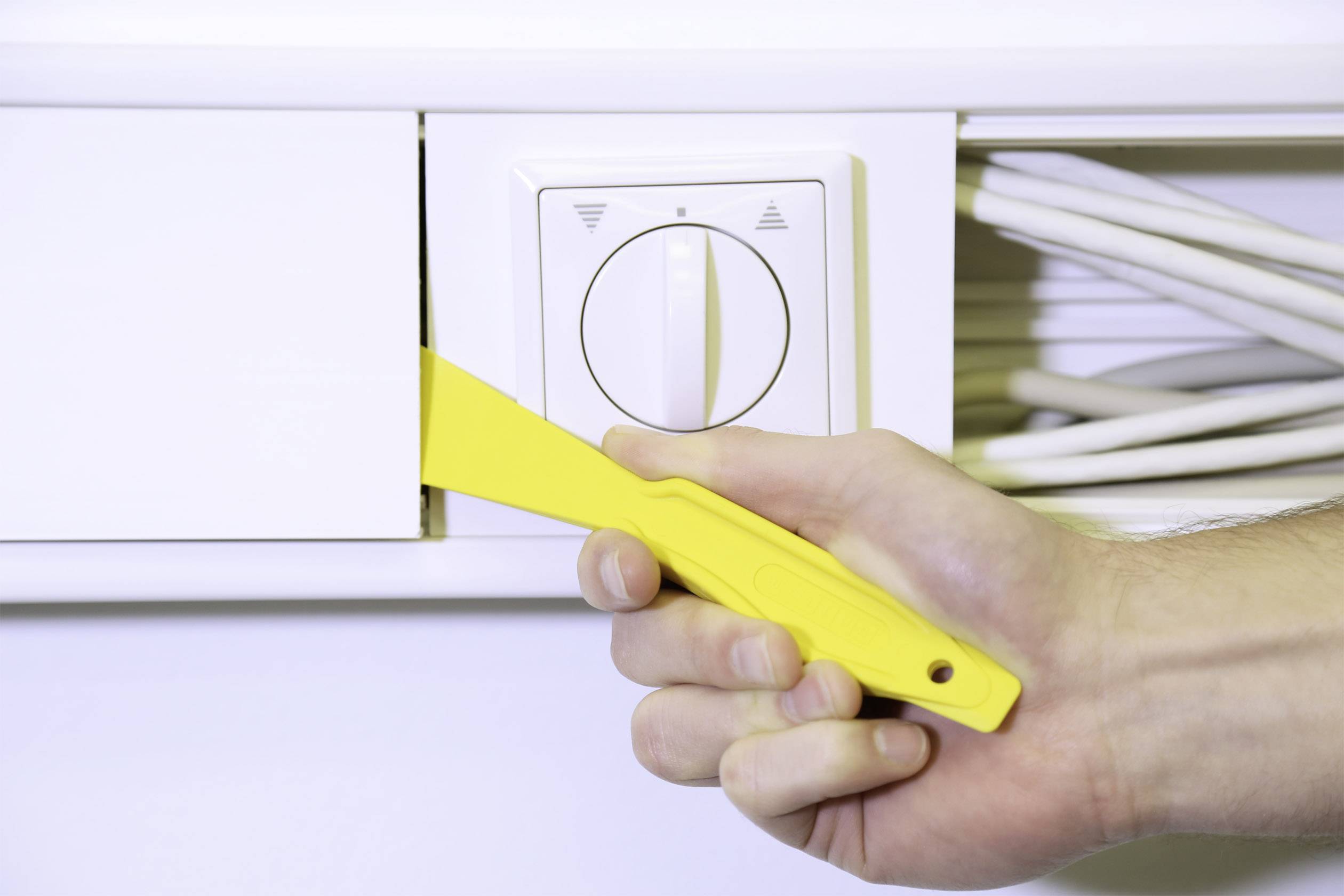 Ręka trzyma żółty plastikowy dźwignik i otwiera białą płytę na powierzchni z kablami, umożliwiając dostęp do urządzeń.