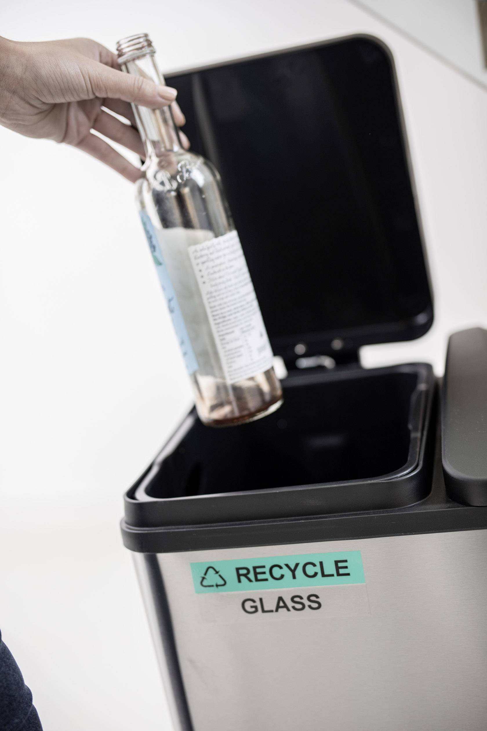 Osoba wrzuca pustą butelkę szklaną do kosza na śmieci z napisem "Recycle Glass".
