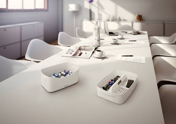 Sala konferencyjna z białymi krzesłami. Dwa organizery z długopisami i artykułami biurowymi na stole. Jasne okna w tle.