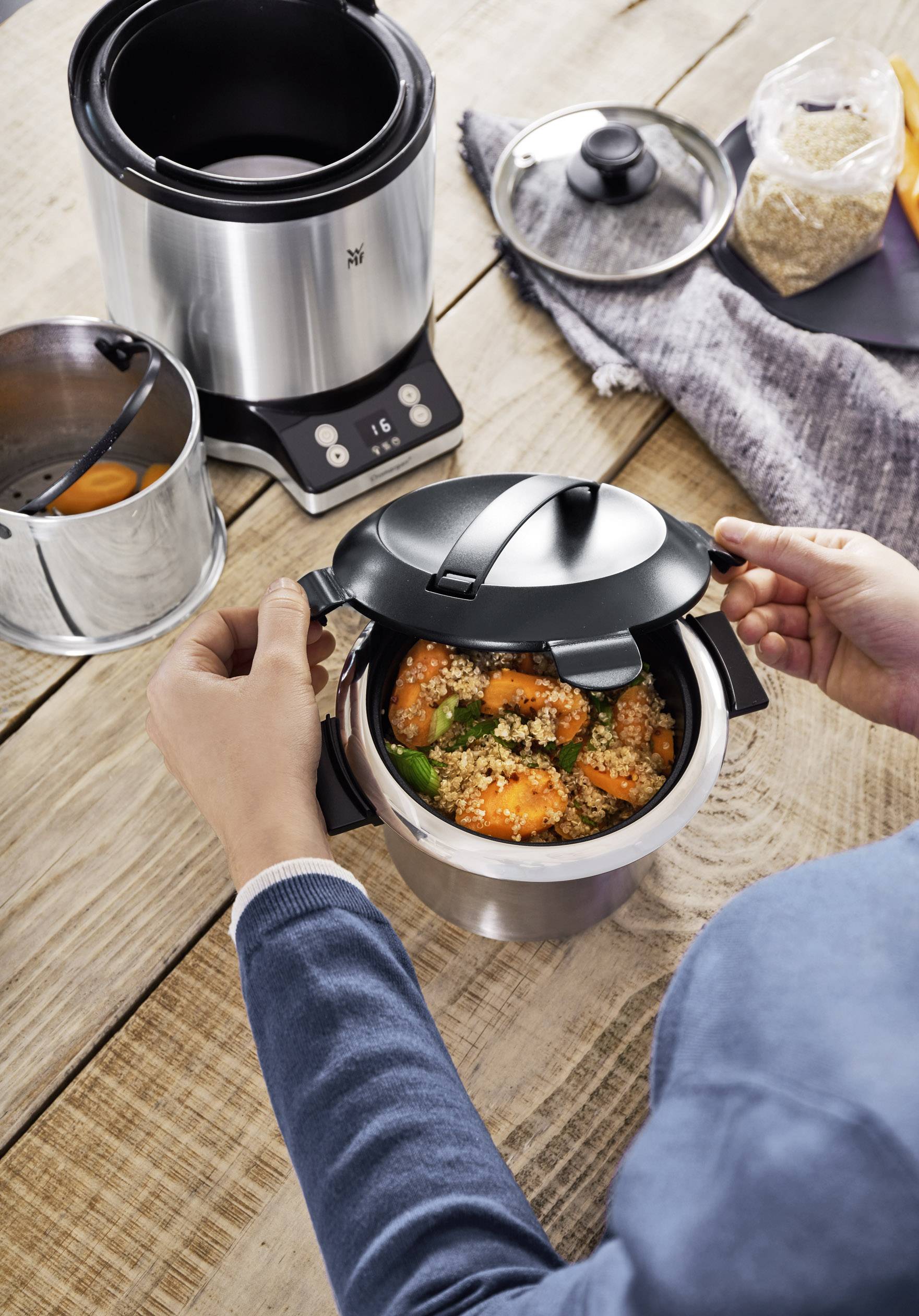 Osoba otwiera garnek ciśnieniowy na drewnianym stole. Garnek zawiera ugotowane warzywa. W tle stoi kolejny garnek.