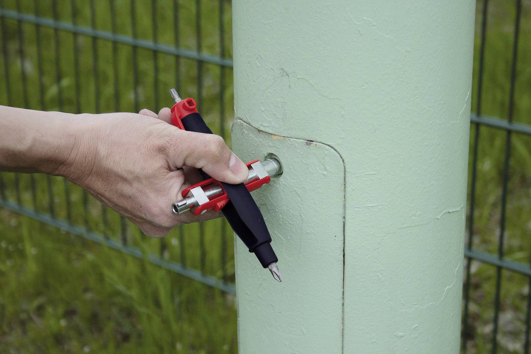 Ręka trzyma multitool, aby przymocować śrubę do słupka ogrodzeniowego. Na tle zielone ogrodzenie.