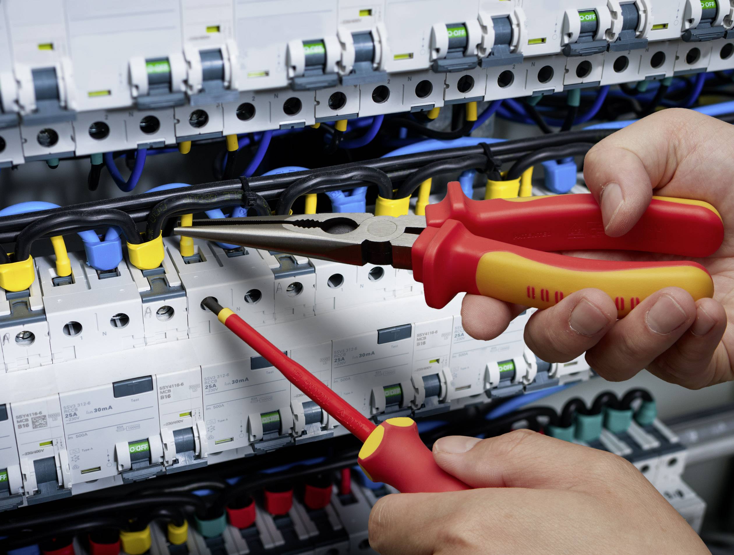 Elektryk używa szczypiec i śrubokrętów podczas pracy przy rozdzielnicy elektrycznej z wieloma obwodami.