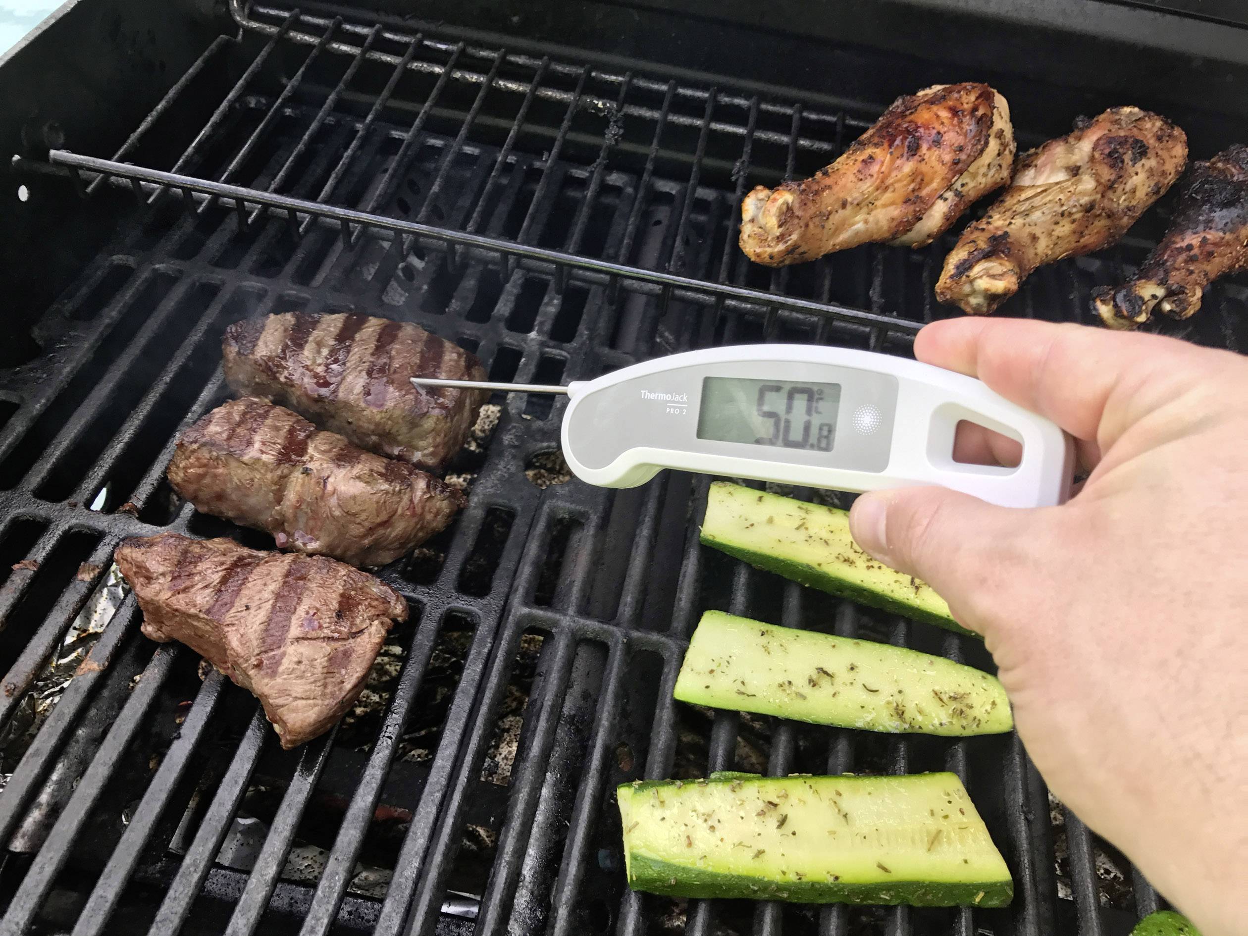 Jedna ręka trzyma termometr do mięsa, który wskazuje temperaturę grillowanego mięsa na 50,8 stopni Celsjusza. Obok leżą kurczak i cukinia.