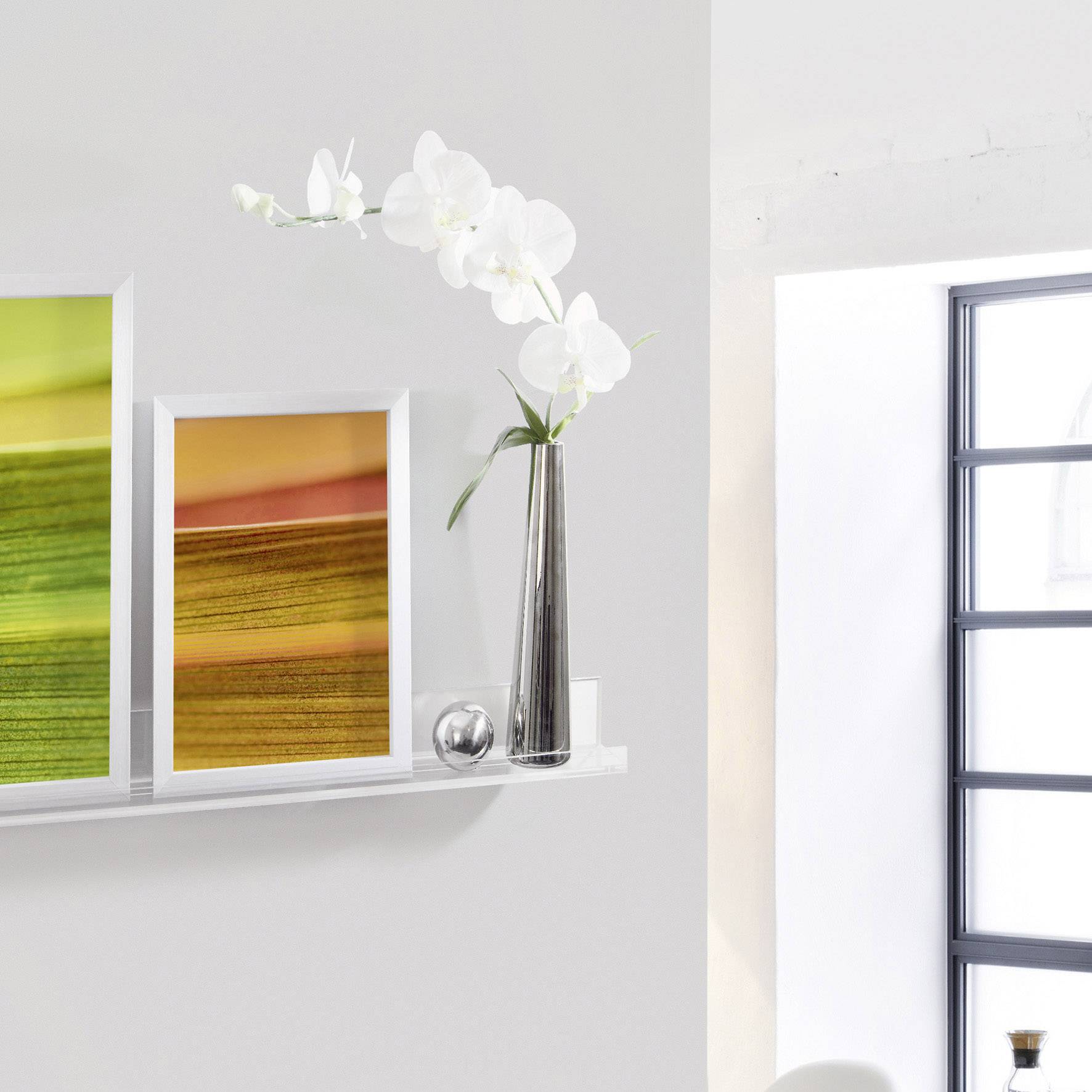 Białe ściany z regałem: Dwa kolorowe obrazy i wazon z białymi orchideami. Jasne światło słoneczne pada na scenę.