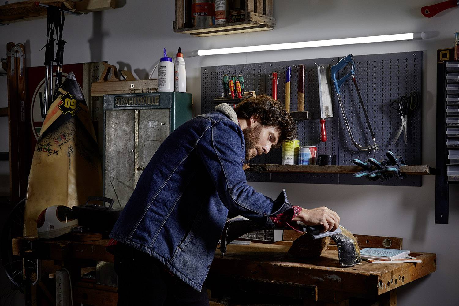 Mężczyzna obrabia kawałek drewna papierem ściernym w warsztacie. W tle widać narzędzia i półki na ścianie.