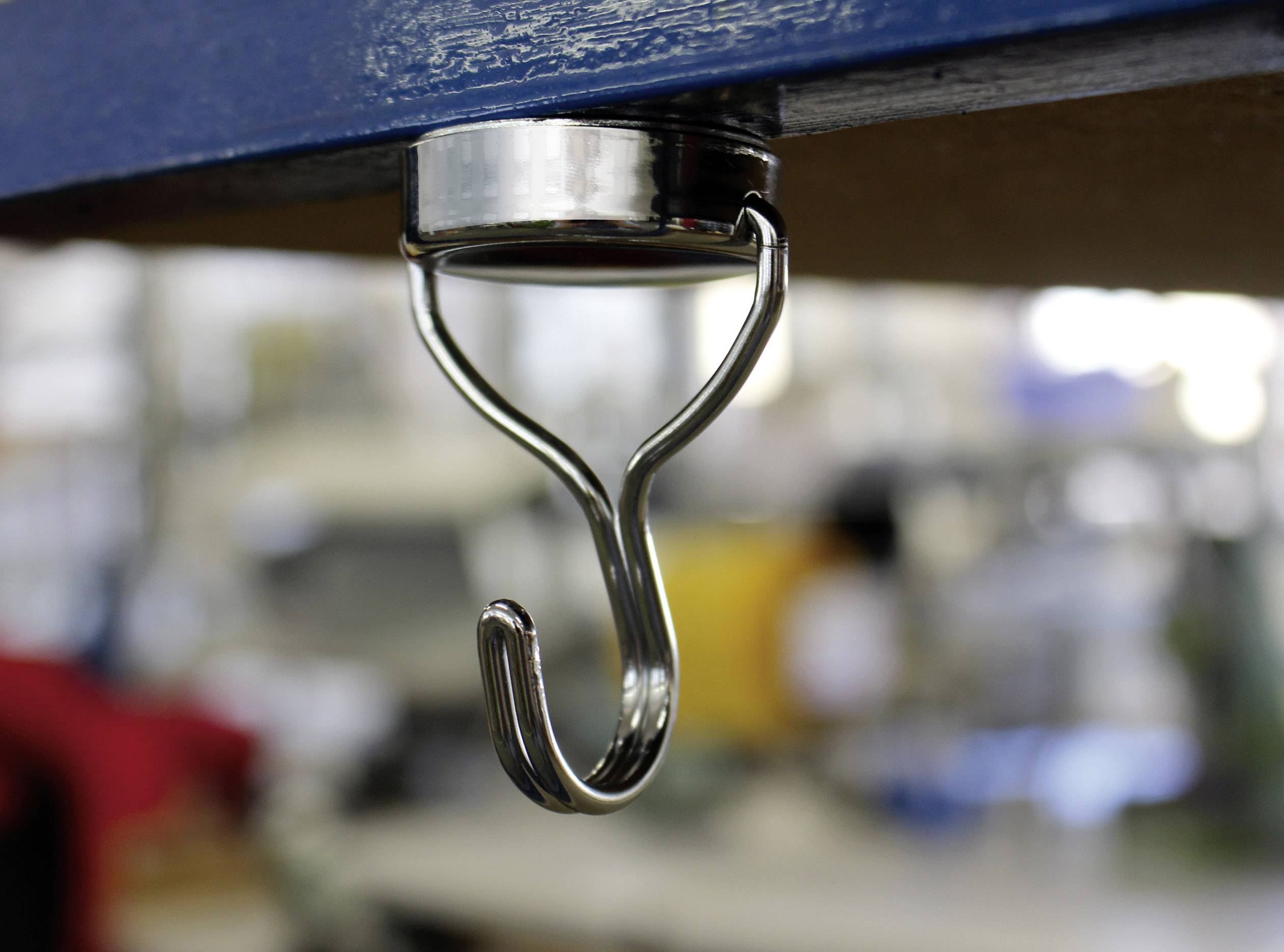 Metalowy hak pod stołem, przeznaczony do przechowywania lub zawieszania przedmiotów w środowisku pracy. Rozmyte tło.