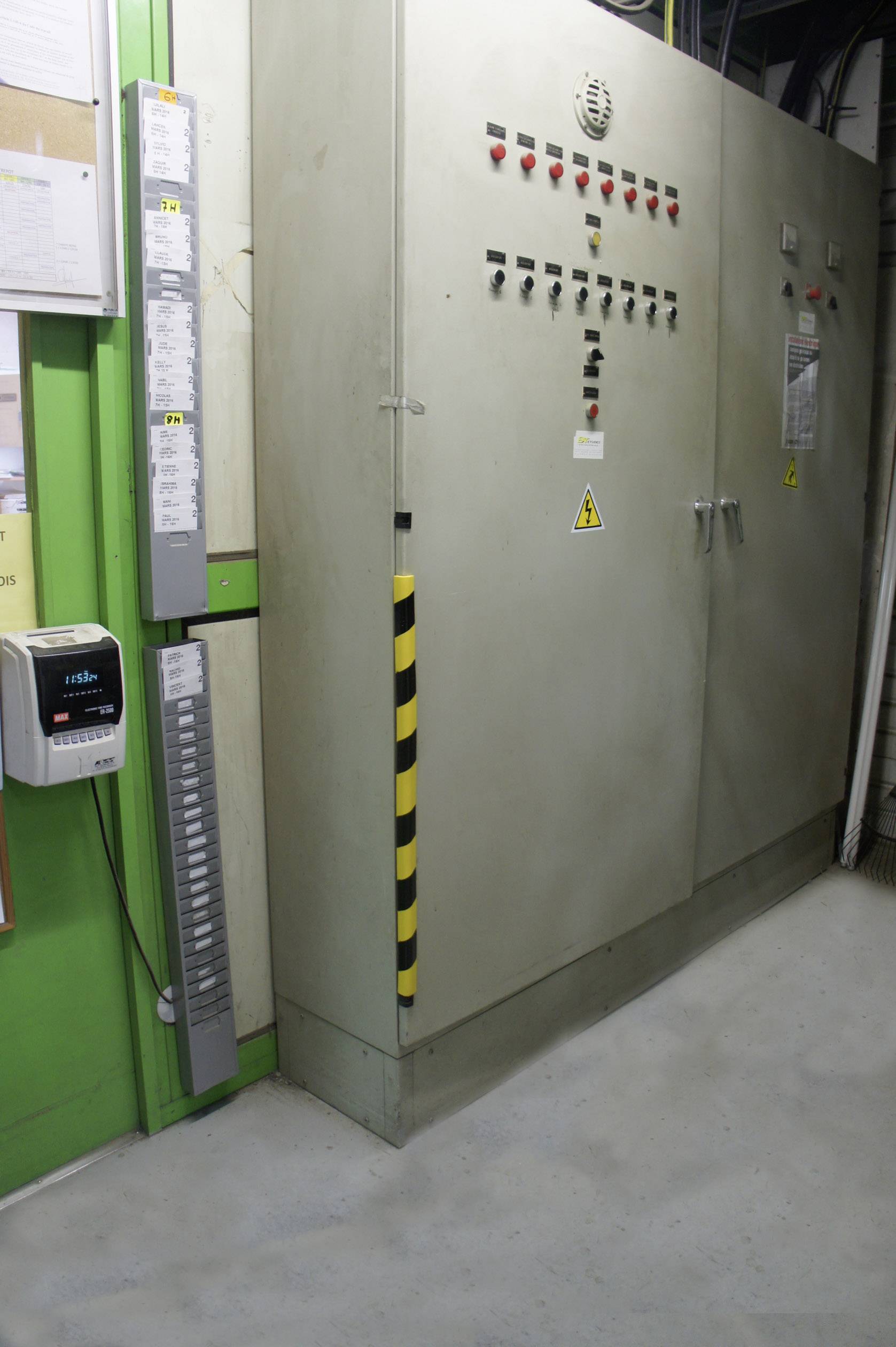 Duży rozdzielnik elektryczny z tablicą ostrzegawczą i wskaźnikami kontrolnymi w środowisku przemysłowym, otoczony zieloną ścianą i dokumentami.