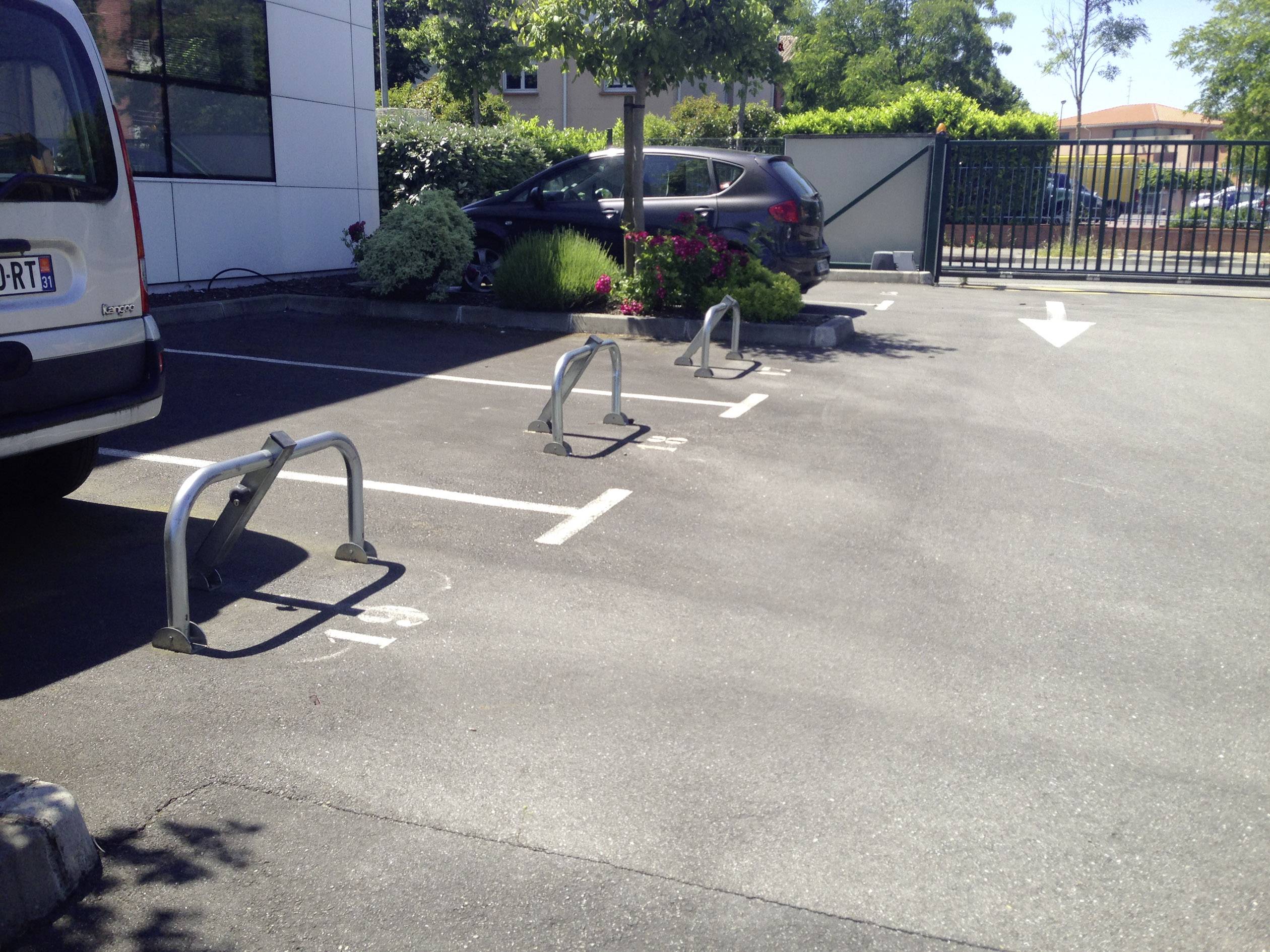 Parking z trzema pustymi miejscami parkingymi zabezpieczonymi metalowymi barierkami. Jeden samochód stoi zaparkowany w tle.