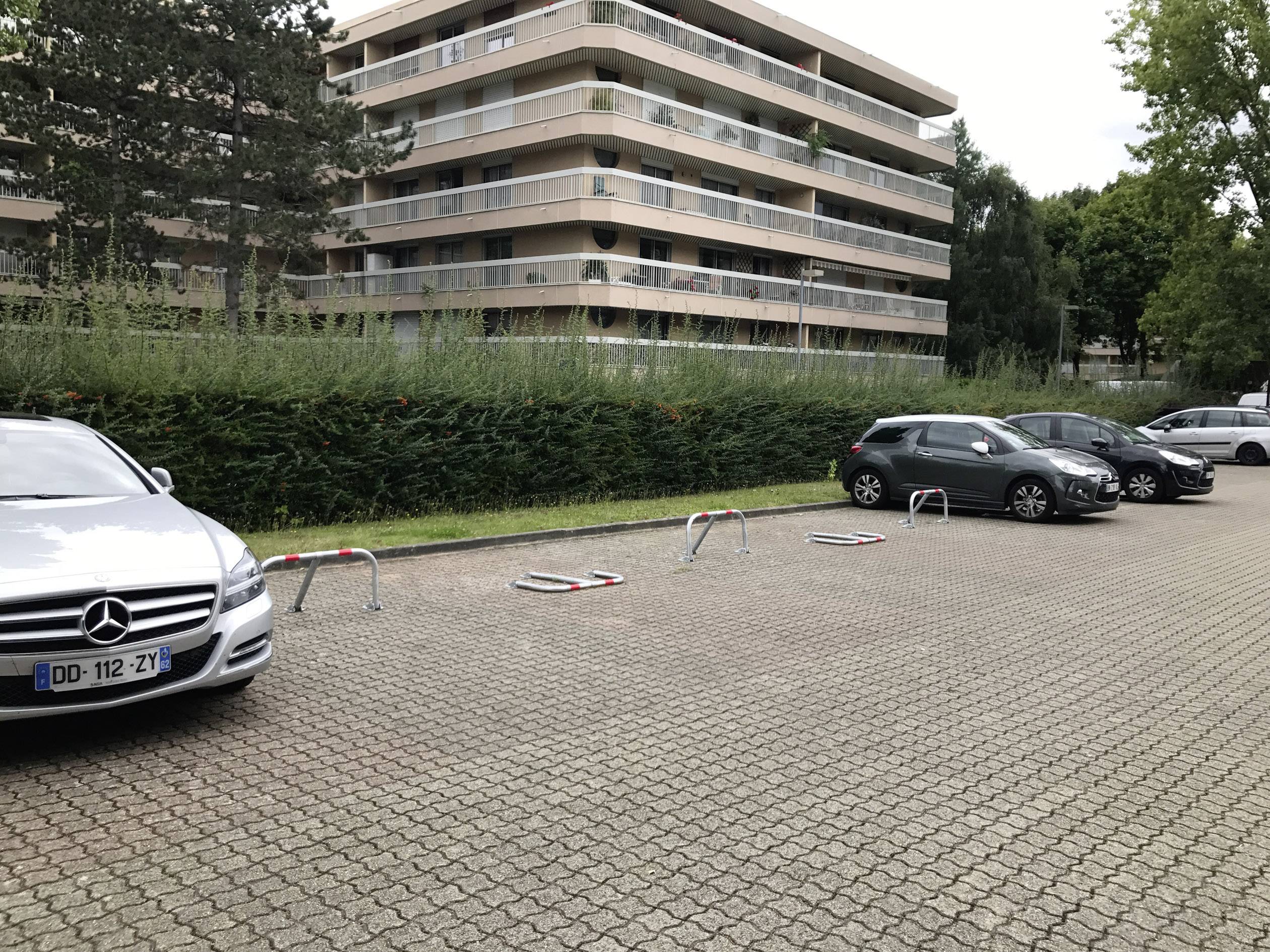 Kilka samochodów parkuje na wybrukowanym parkingu przed budynkiem mieszkalnym. Na parkingu zainstalowane są słupki parkingowe.