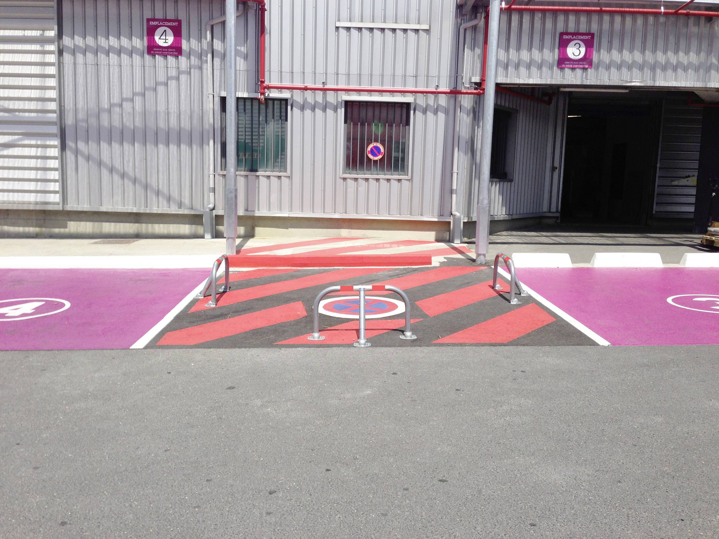 Czerwono-biało oznaczony obszar przed budynkiem wskazuje na zakaz parkowania. Obok widoczne są różowe miejsca parkingowe z numeracją.