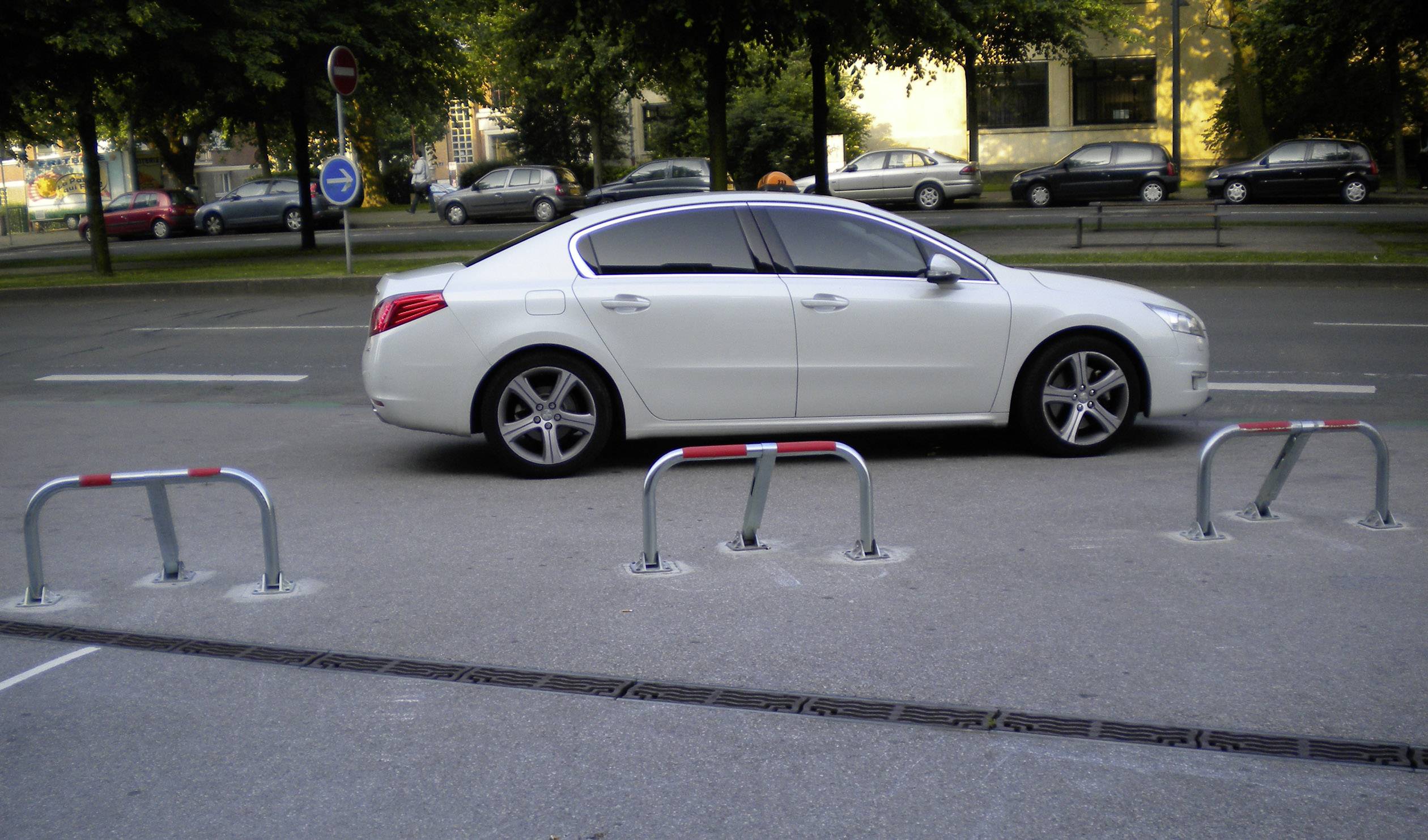 Białe auto jest nieprawidłowo zaparkowane wzdłuż ulicy między trzema słupkami na pustym parkingu. W tle stoją liczne samochody.