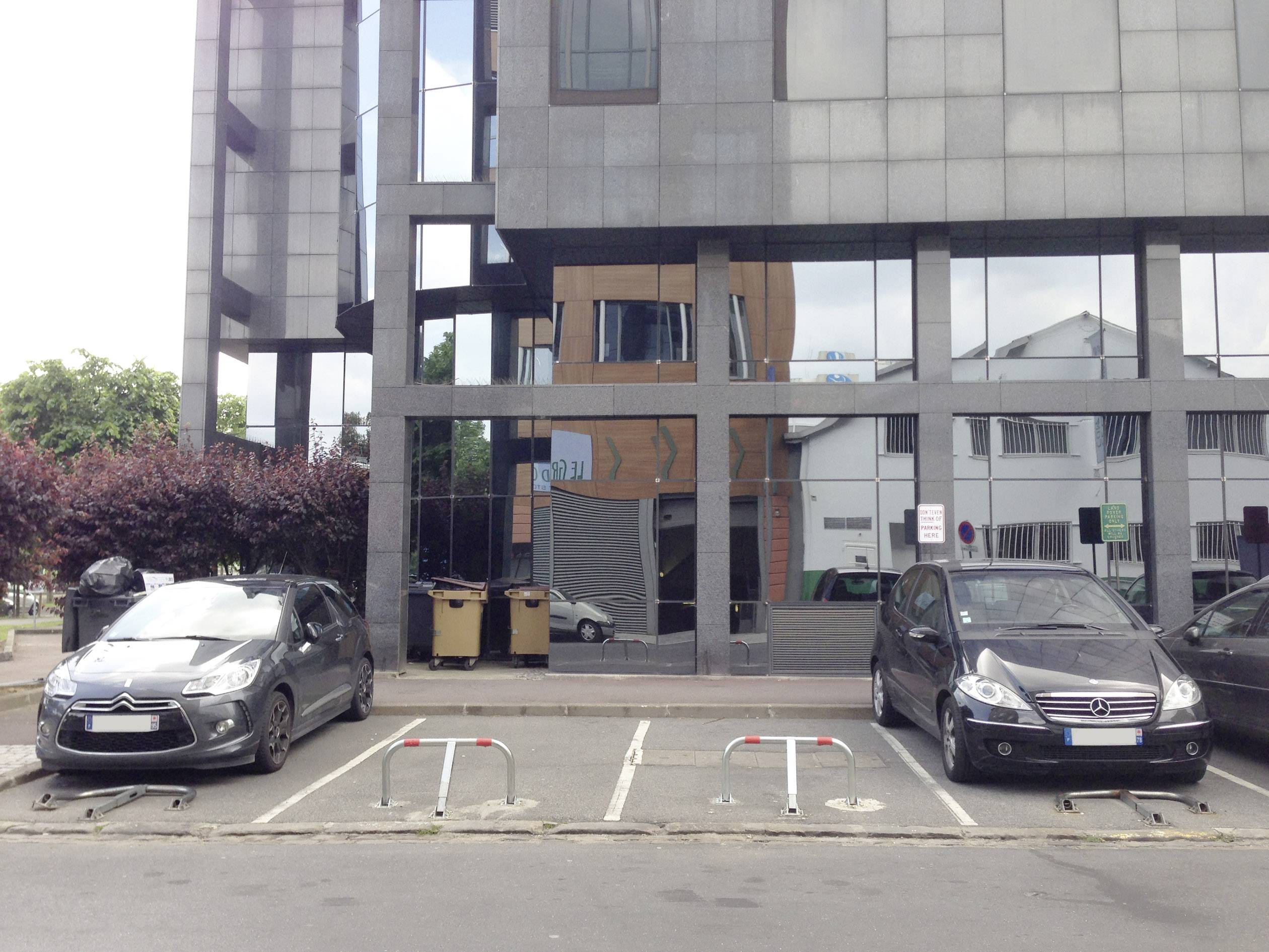 Dwa puste miejsca parkingowe przed budynkiem, otoczone zaparkowanymi samochodami. Tablica informuje o warunkach parkowania.