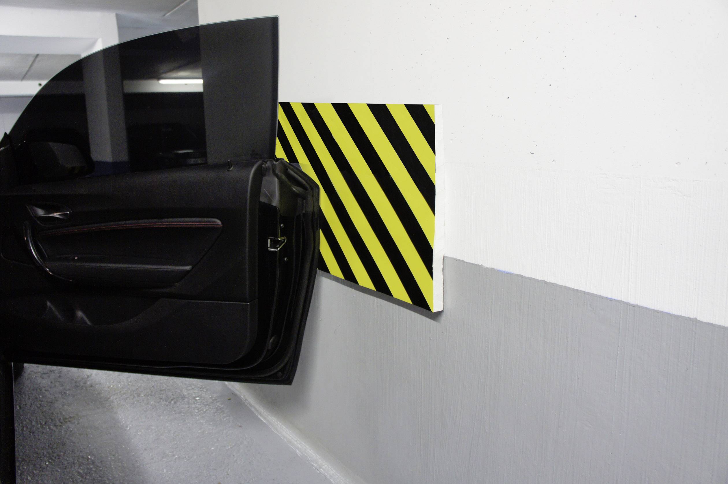 Drzwi samochodu otwierają się w parkingu przy ścianie oznaczonej czarno-żółtymi pasami bezpieczeństwa, które mają zapobiegać uszkodzeniom.