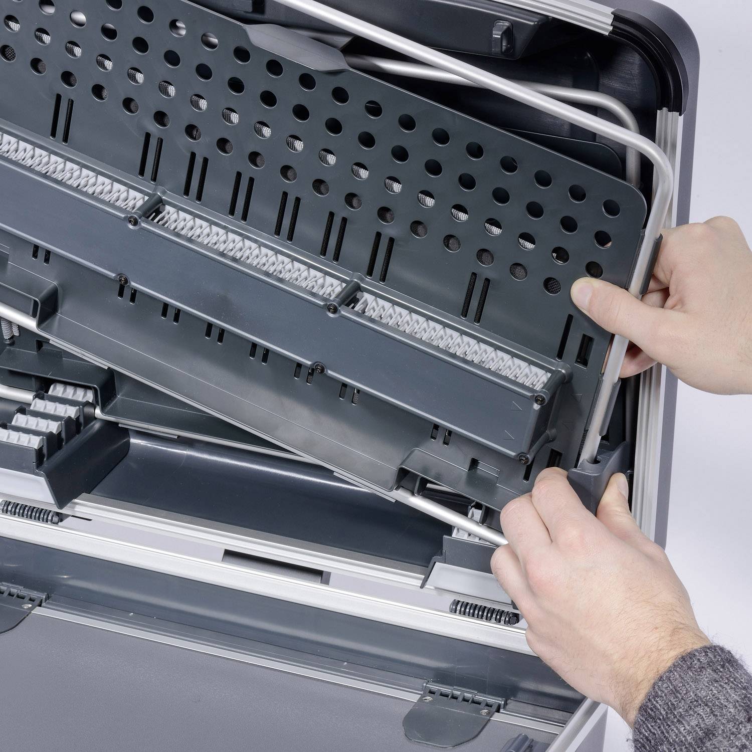 Dwie dłonie otwierają szary kufer z wysuwaną rączką. Wygląda on na odpowiedni do przechowywania narzędzi lub sprzętu technicznego.