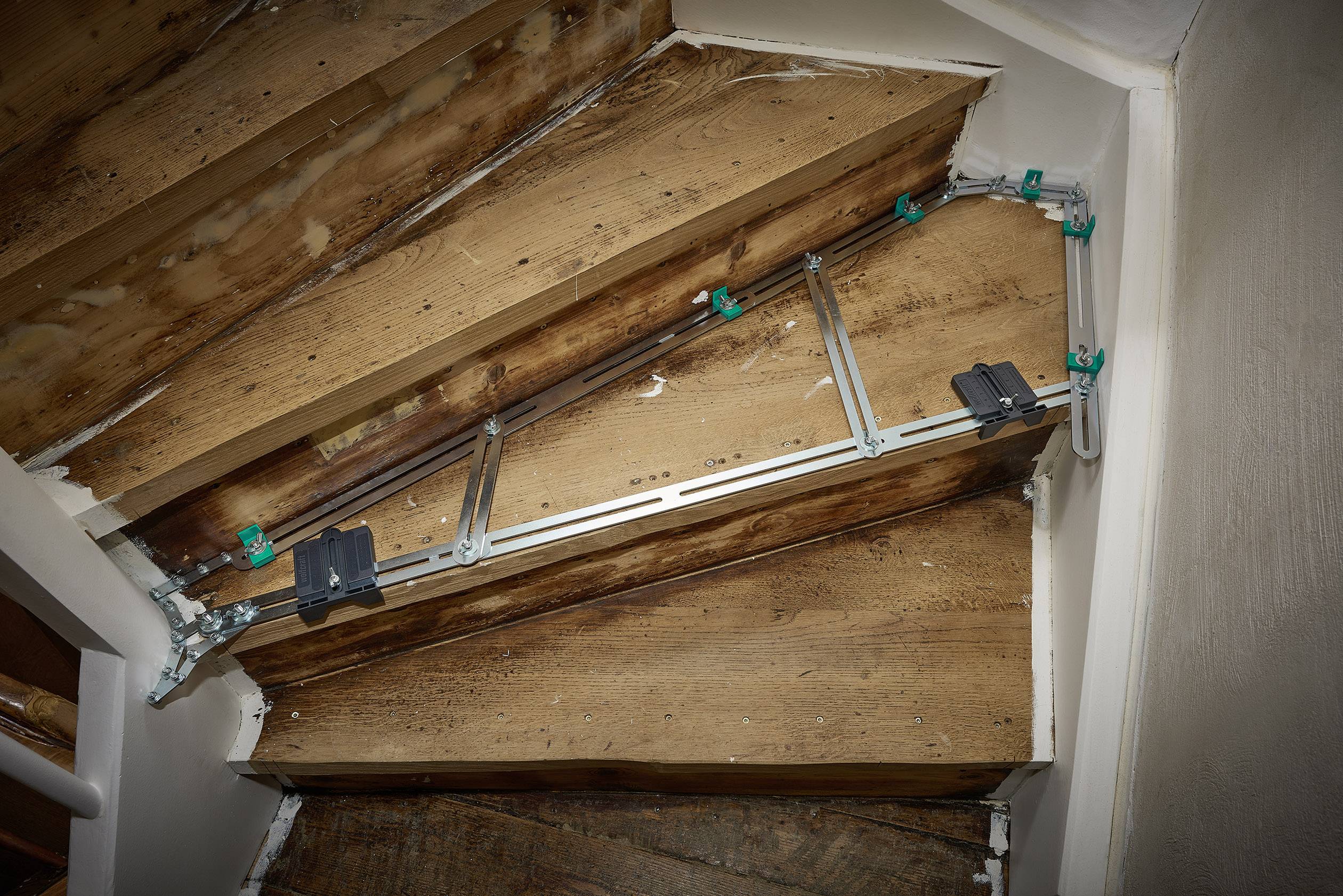 System schodowy zamontowany jest na pochyłej drewnianej konstrukcji schodów, prezentując metalowe prowadnice z zielonymi uchwytami.