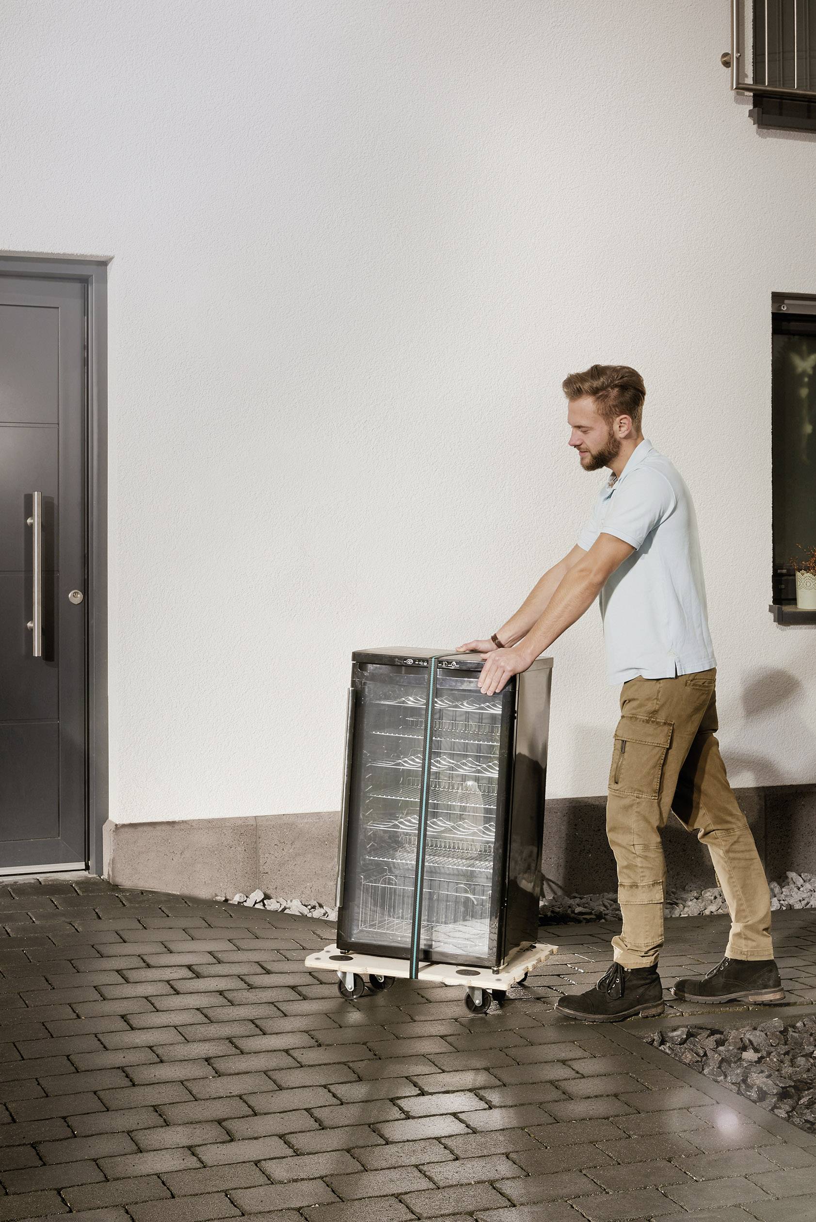 Osoba przesuwa czarną lodówkę na wózku transportowym przez wybrukowaną alejkę w pobliżu domu.