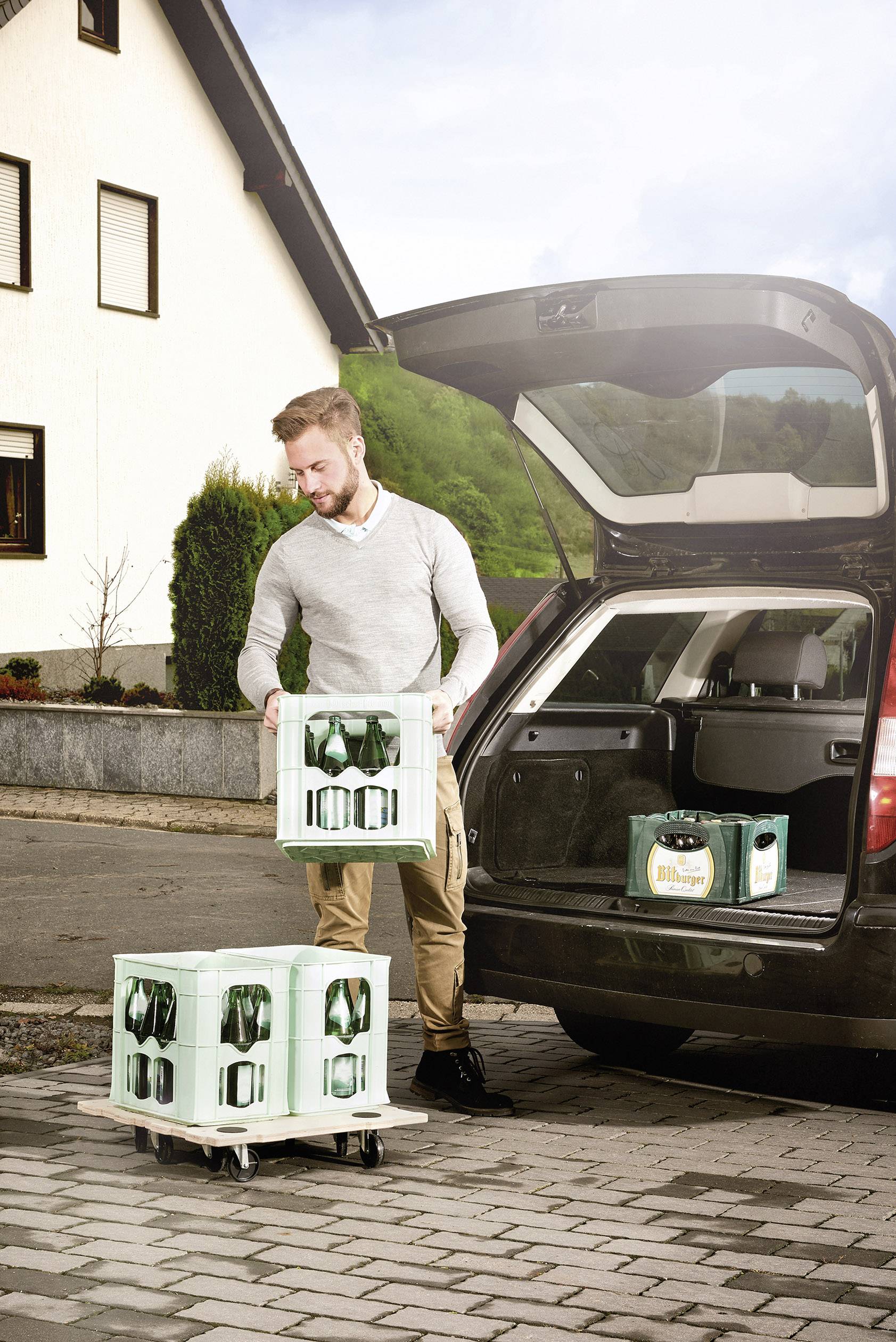 Mężczyzna wyładowuje skrzynie z napojami z bagażnika samochodu na parkingu. W tle widać dom mieszkalny.
