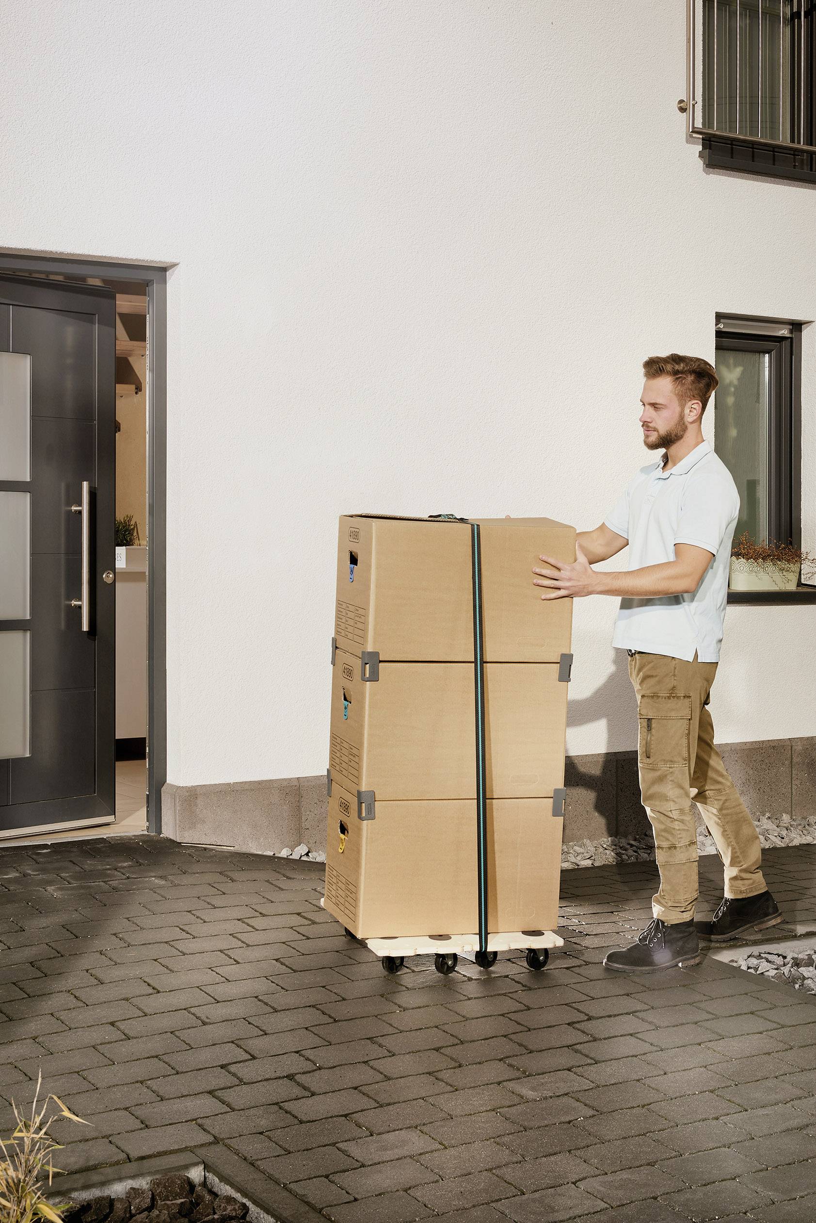 Mężczyzna przewozi trzy ułożone kartonowe pudła na wózku transportowym przed nowoczesnym wejściem do budynku.