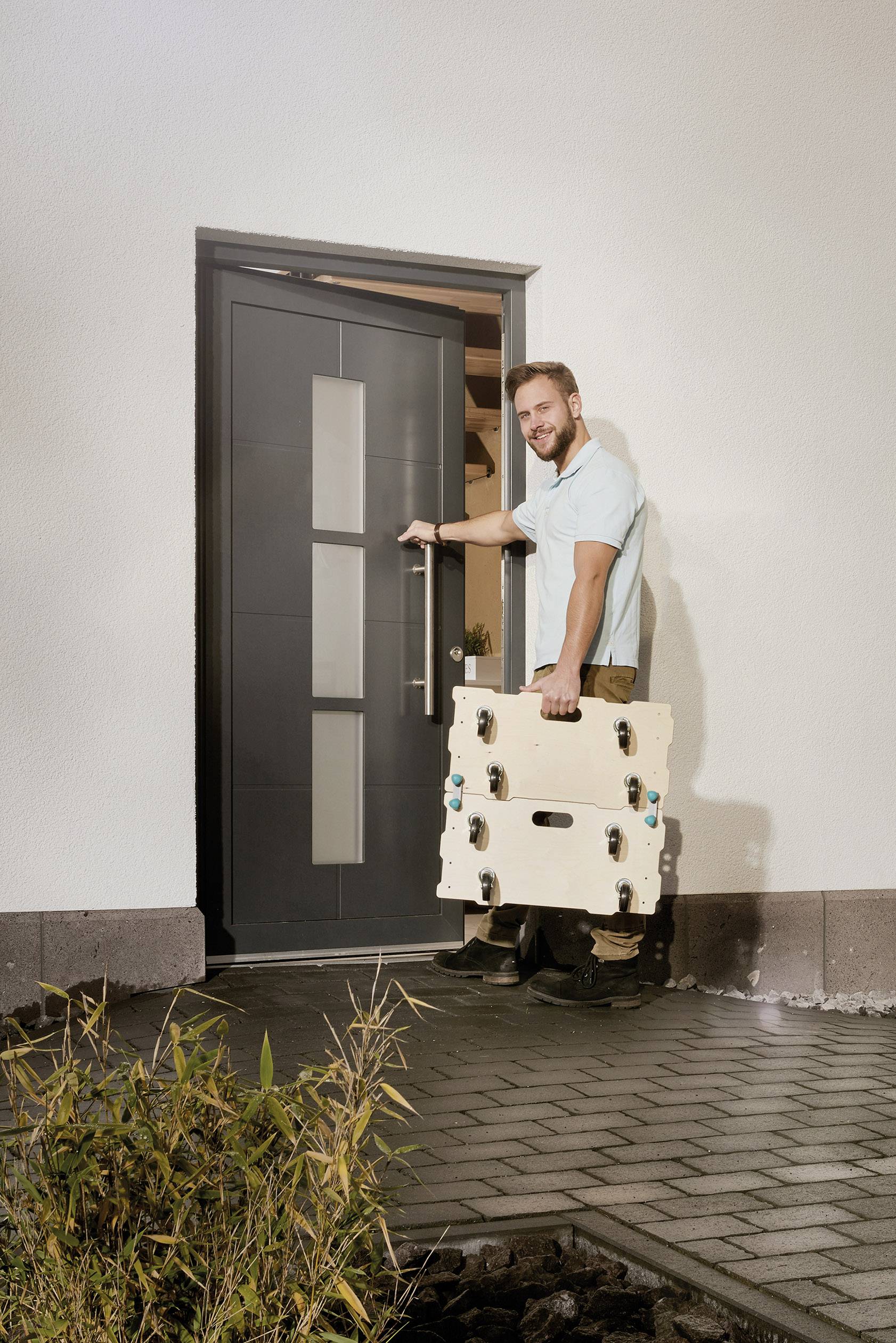 Mężczyzna otwiera drzwi wejściowe i trzyma duży, beżowy kufer na kółkach, gotowy, aby wejść do środka. Na pierwszym planie widoczne są rośliny.