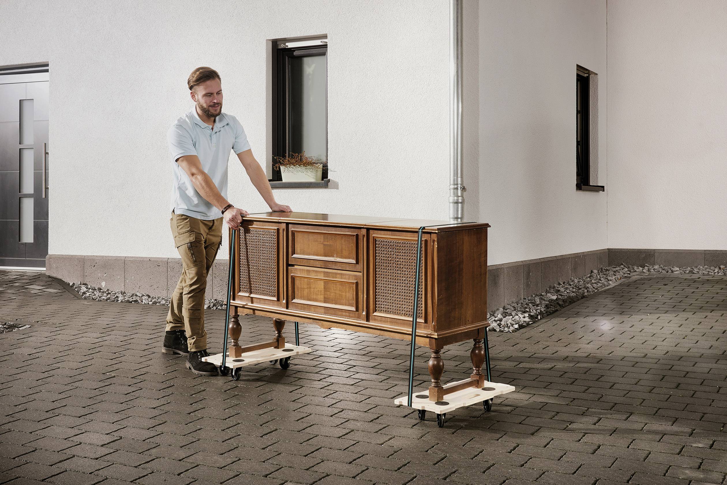 Osoba przemieszcza zabytkowy mebel na wózkach transportowych przez brukowany dziedziniec. Mebel jest zabezpieczony pasami.