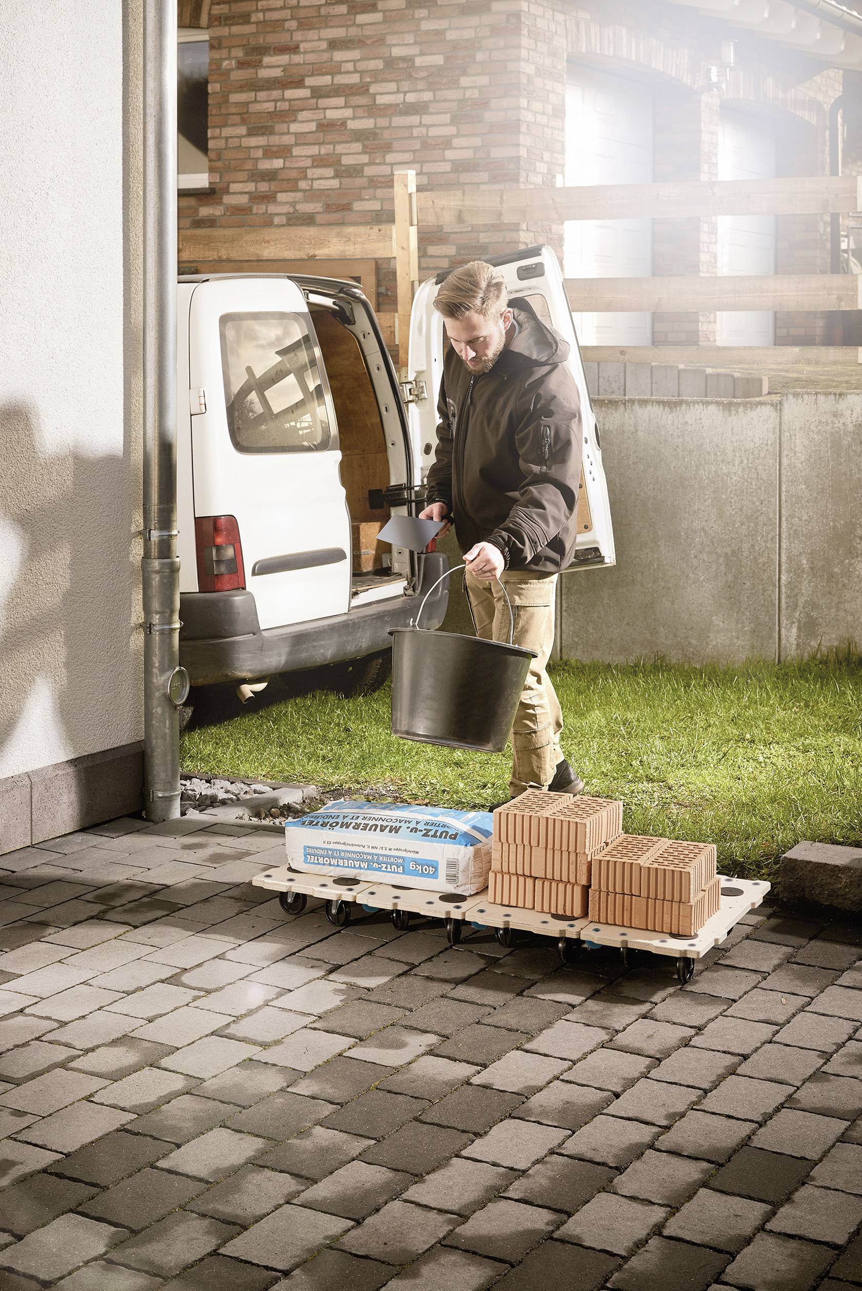 Mężczyzna rozładowuje paletę z cegłami i torbę cementu przed vanem na podwórzu. Trzyma wiadro.