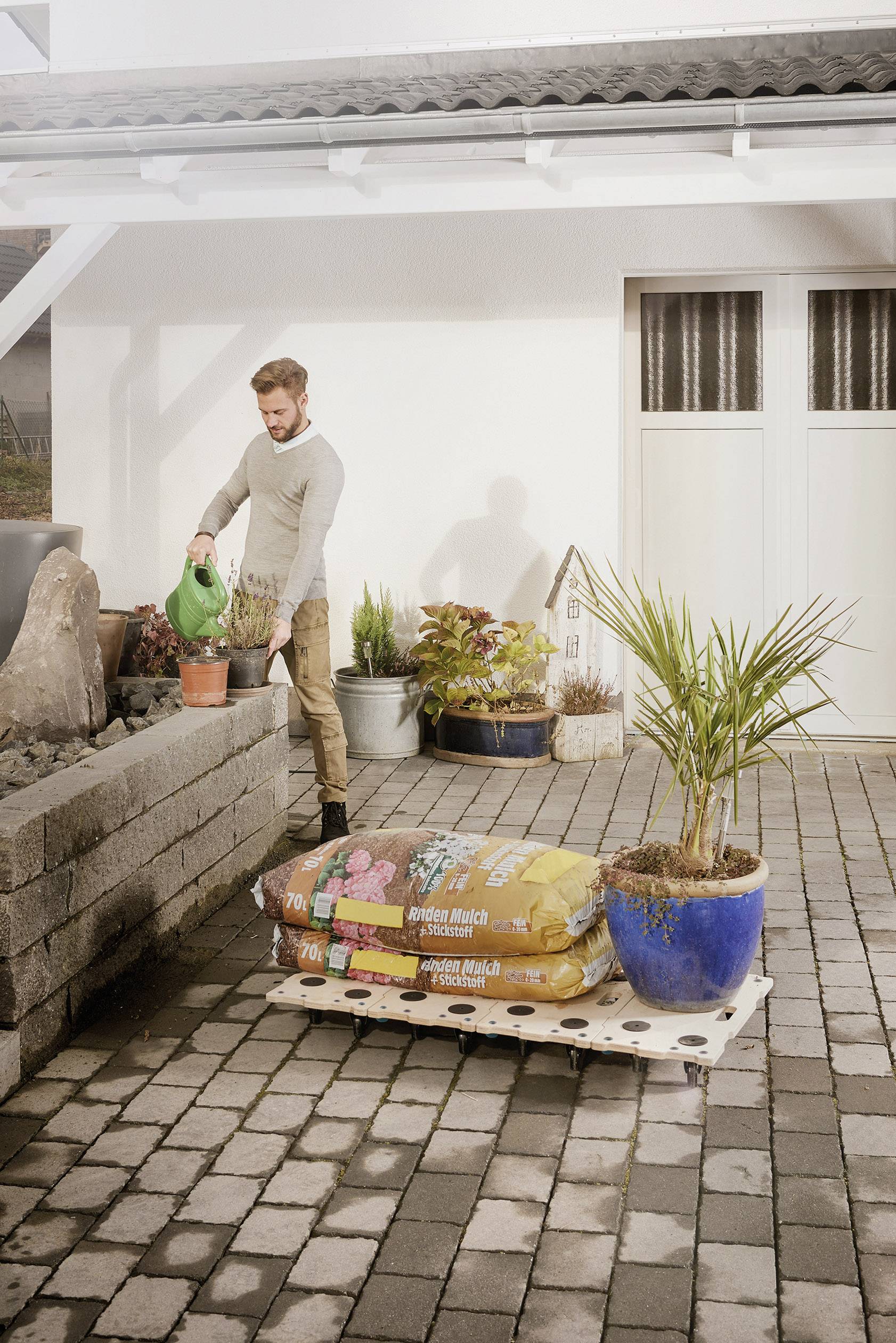 Mężczyzna podlewa rośliny w ogrodzie wyłożonym kostką brukową. Na pierwszym planie stoją worki z ziemią i duży doniczka.