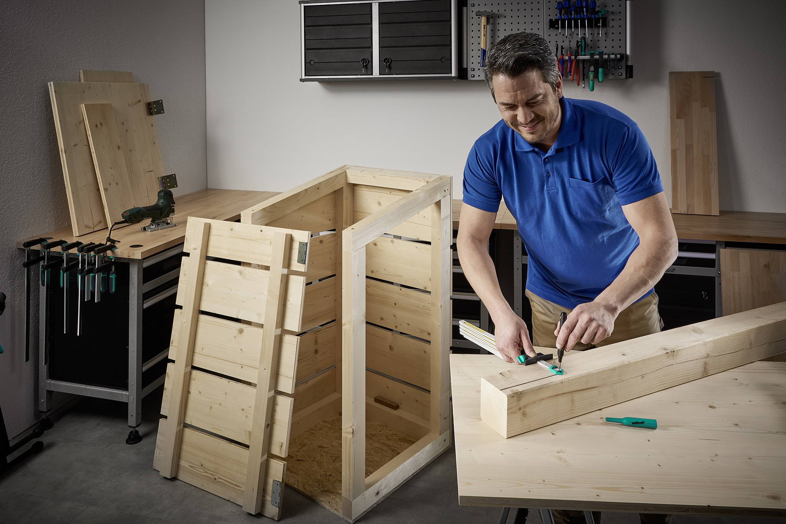 Mężczyzna w niebieskiej koszulce polo montuje drewniane pudło w warsztacie. W tle widoczne są narzędzia i dodatkowe deski drewniane.