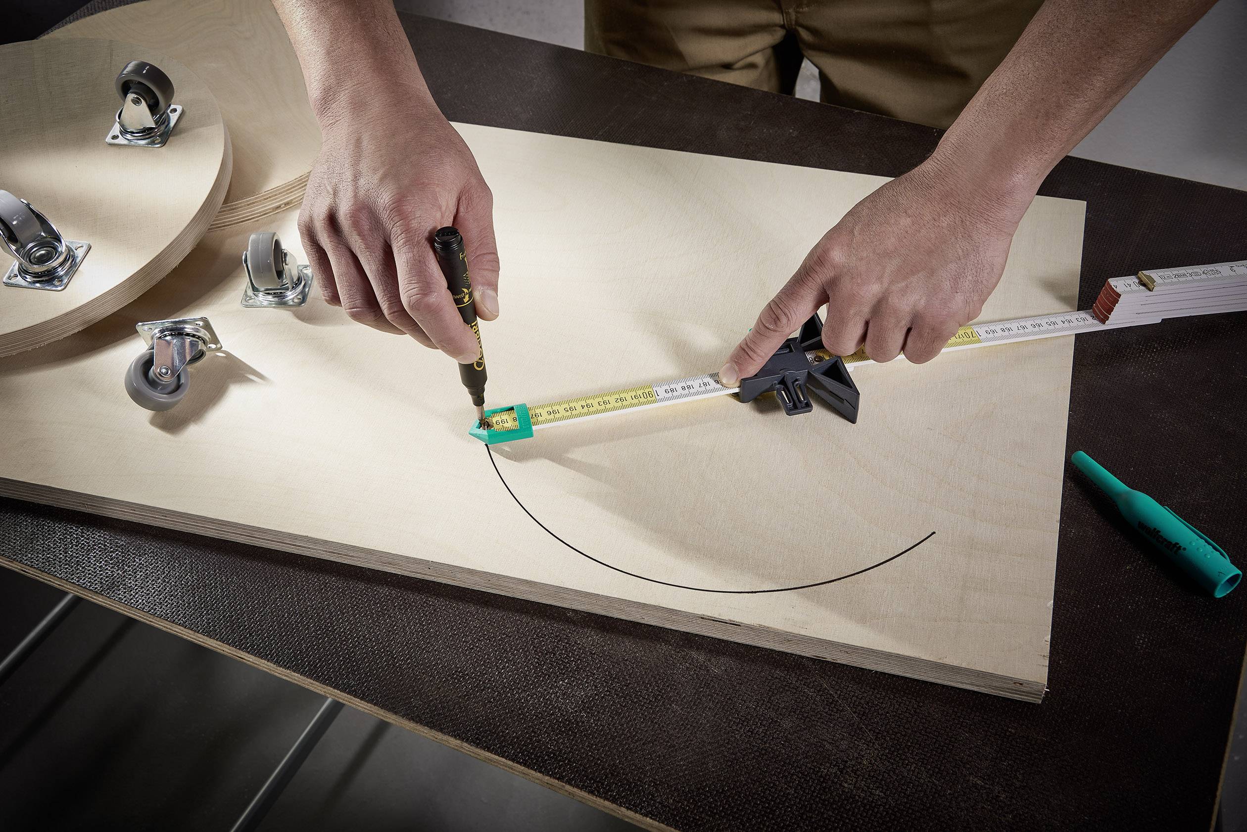 Osoba rysuje koło na desce drewnianej za pomocą miarki i pisaka. W tle widoczne są pojedyncze rolki.