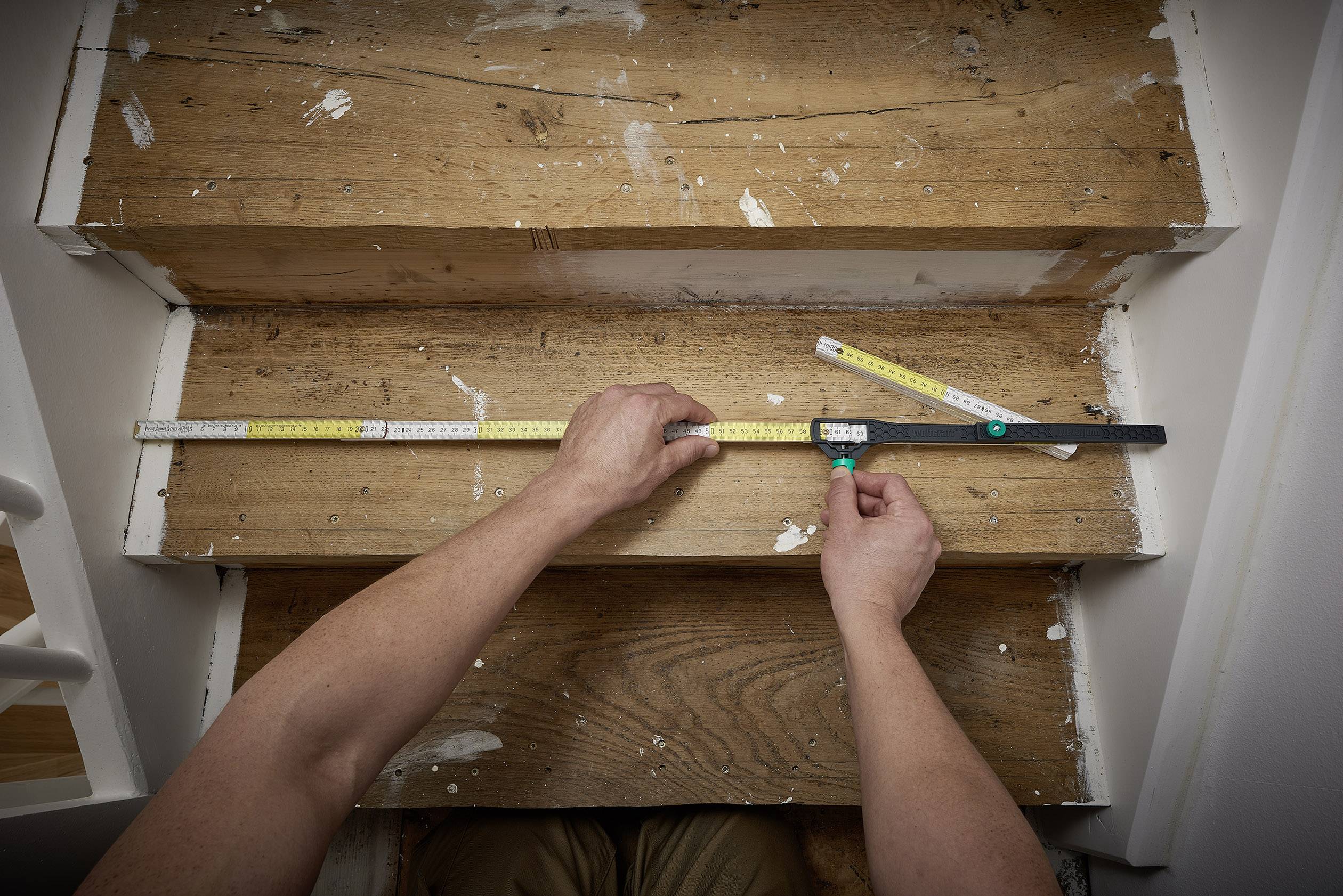 Jedna osoba mierzy szerokość i głębokość drewnianego stopnia schodów za pomocą dwóch taśm mierniczych umieszczonych na stopniu.