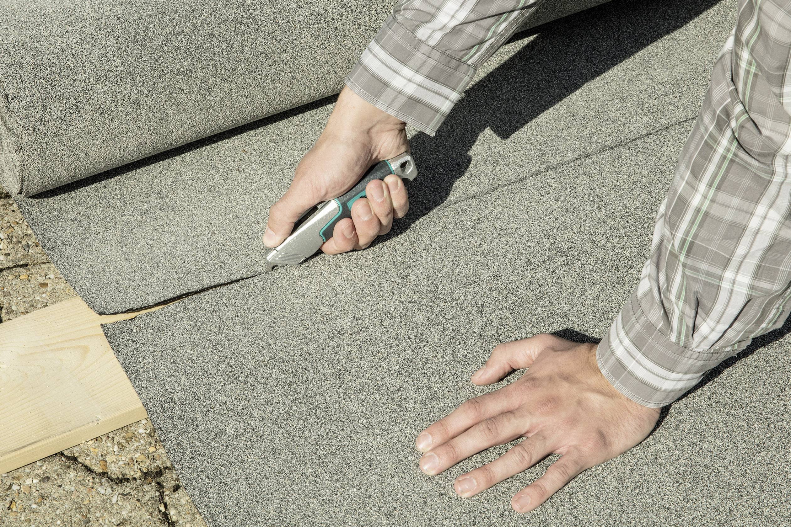 Osoba przycina szary dywan nożem do cięcia na drewnianej desce. Ręce i część narzędzia są widoczne.