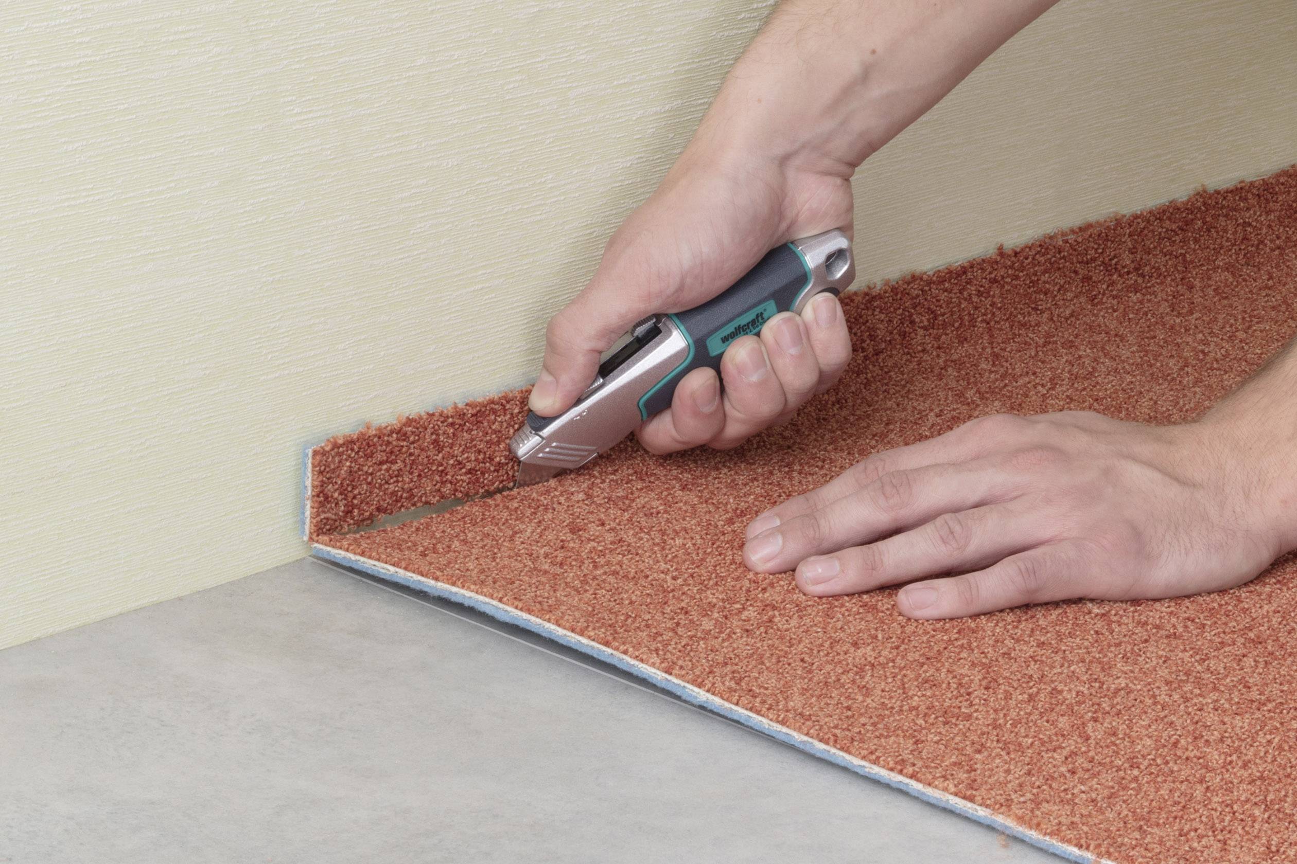 Osoba przycina pomarańczowy dywan wzdłuż ściany za pomocą noża do dywanów, aby dopasować go do pomieszczenia.