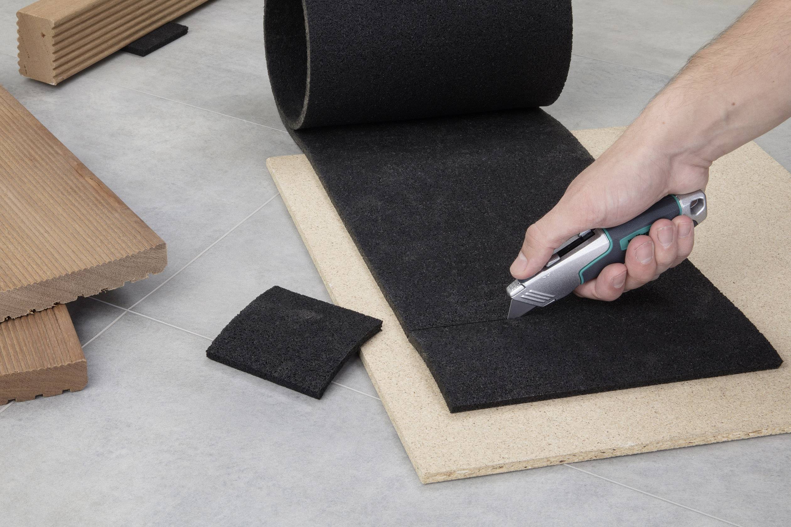 Osoba przecina nożem introligatorskim kawałek czarnego materiału wykładzinowego na drewnianej płycie. W tle widoczne są kawałki drewna.
