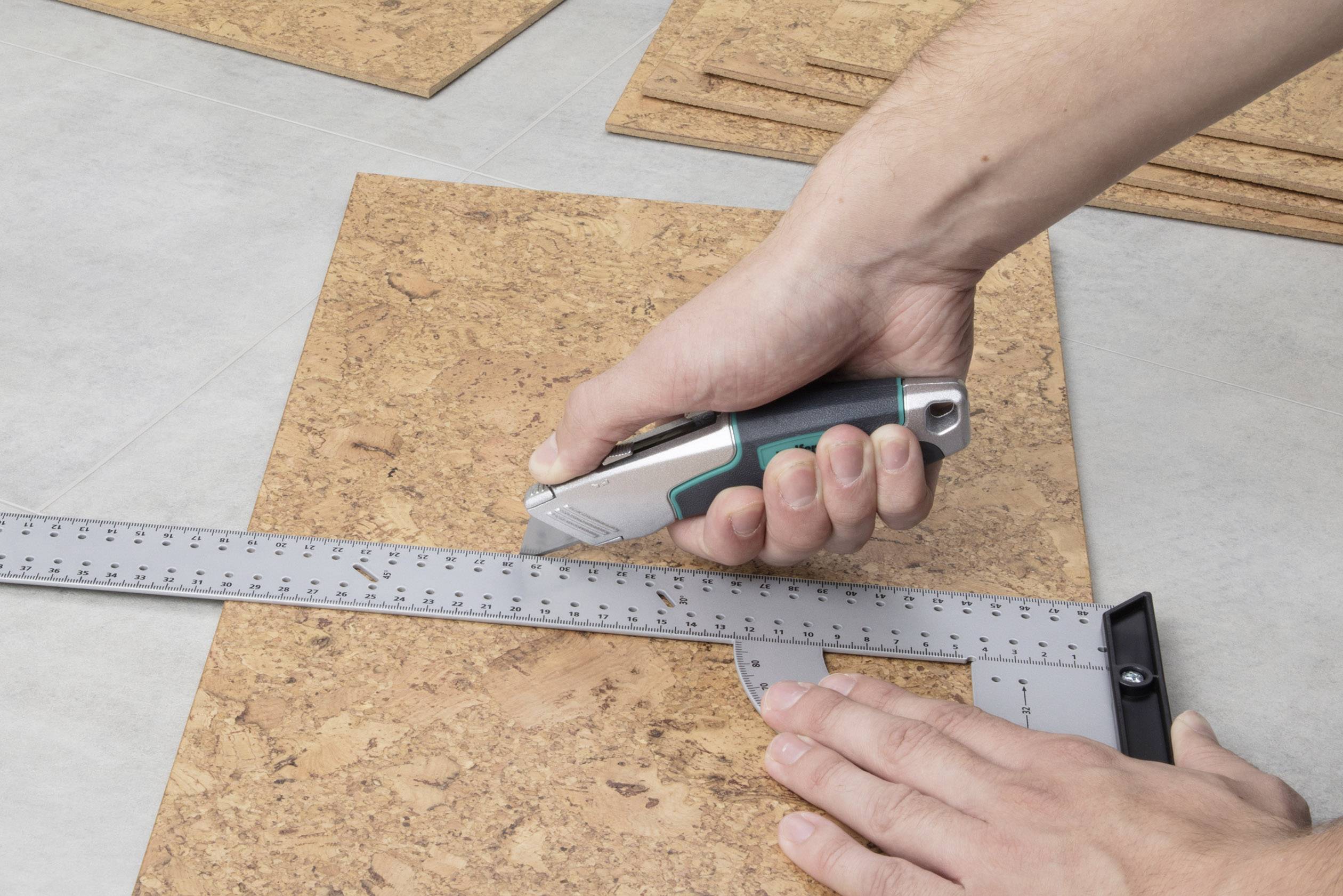 Osoba przecina deskę korkową nożem do cięcia wzdłuż metalowej linijki. Ręce mocno trzymają nóż i linijkę.