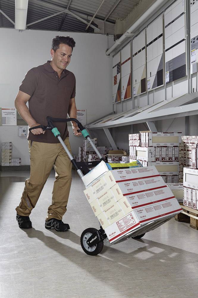 Mężczyzna przewozi ręcznym wózkiem kilka ułożonych jeden na drugim kartonów przez magazyn. Kartony są prostokątne i opisane etykietami.