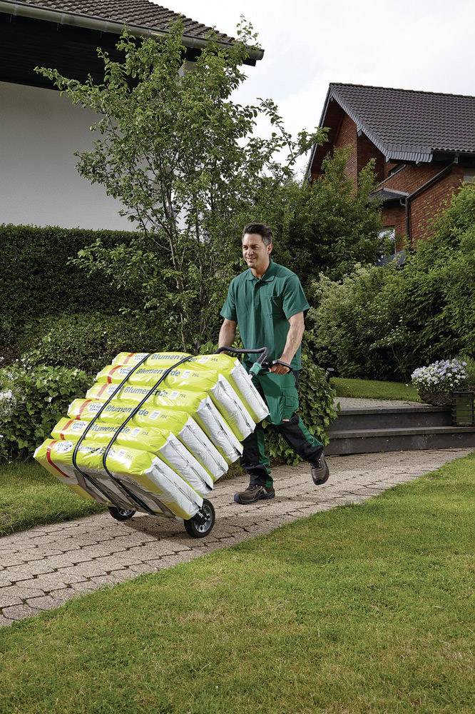 Mężczyzna w zielonej odzieży roboczej popycha wózek ręczny pełen żółtych worków na wybrukowanej drodze przed budynkiem mieszkalnym.