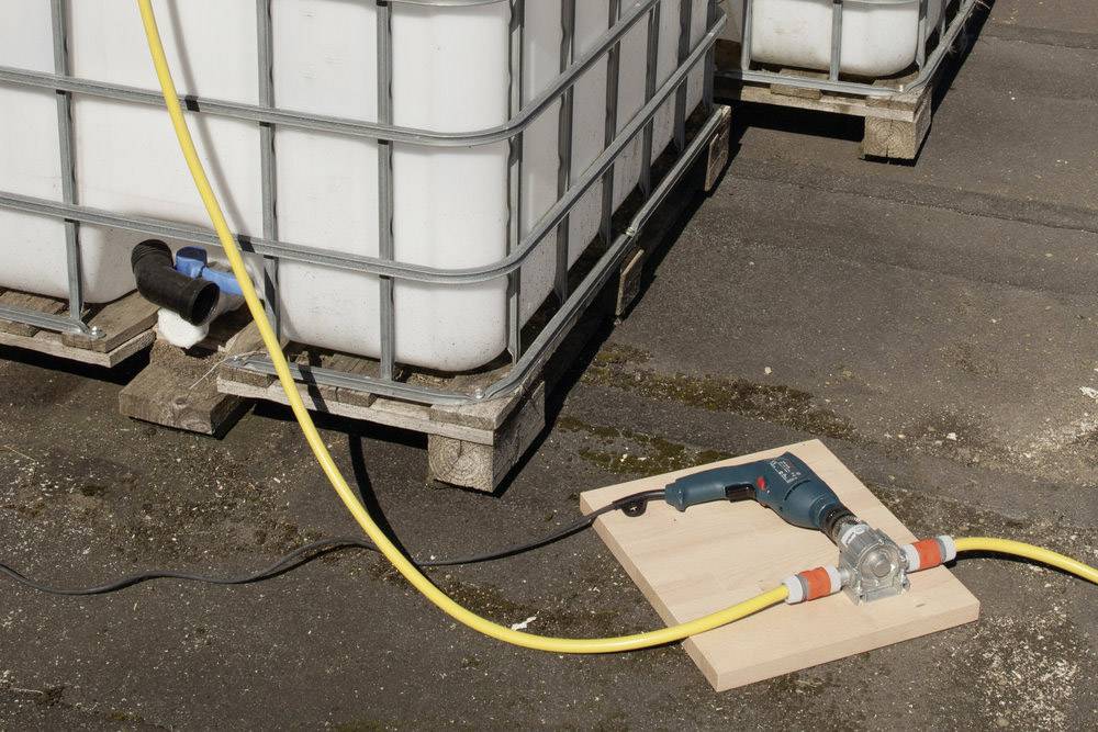 Duży zbiornik na wodę stoi na palecie. Żółty wąż jest podłączony do zbiornika i prowadzi do urządzenia elektrycznego umieszczonego na desce.