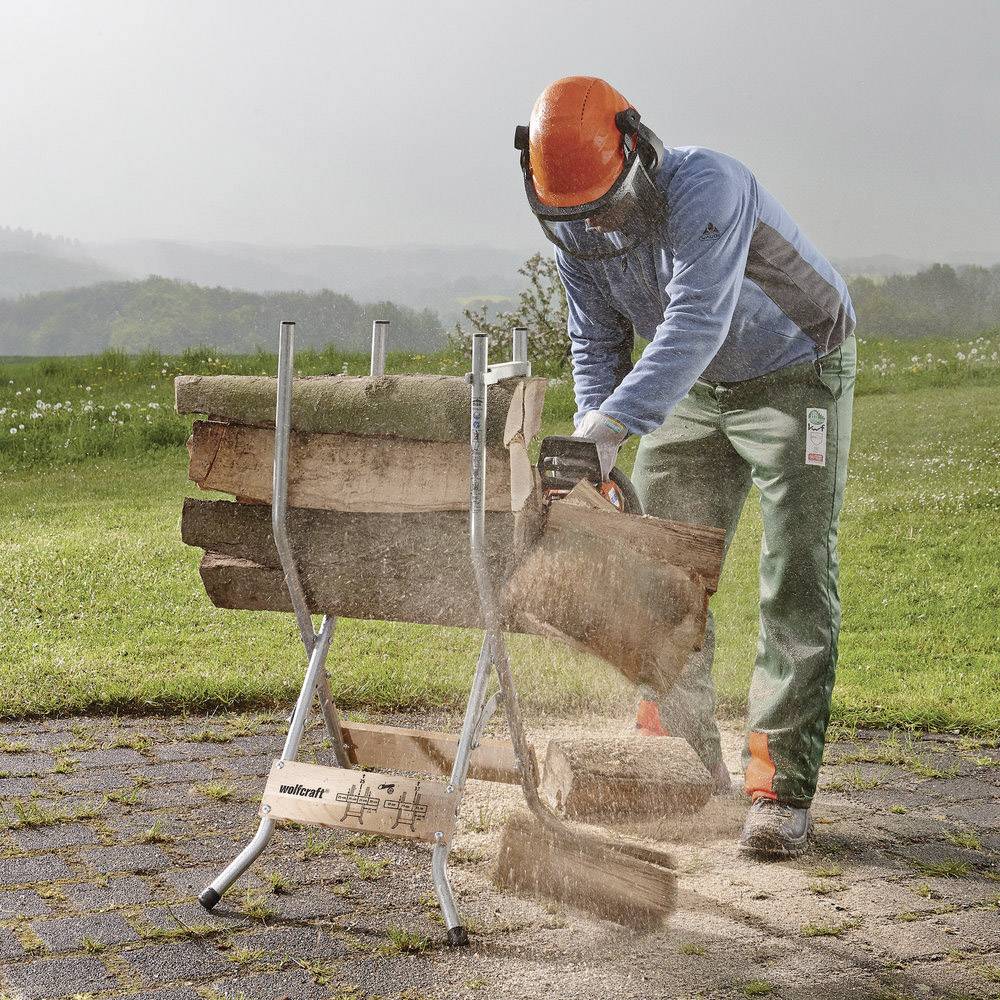 Osoba w odzieży ochronnej piłuje drewno pilarką spalinową na stojaku na świeżym powietrzu. Wióry drewniane unoszą się w powietrzu.