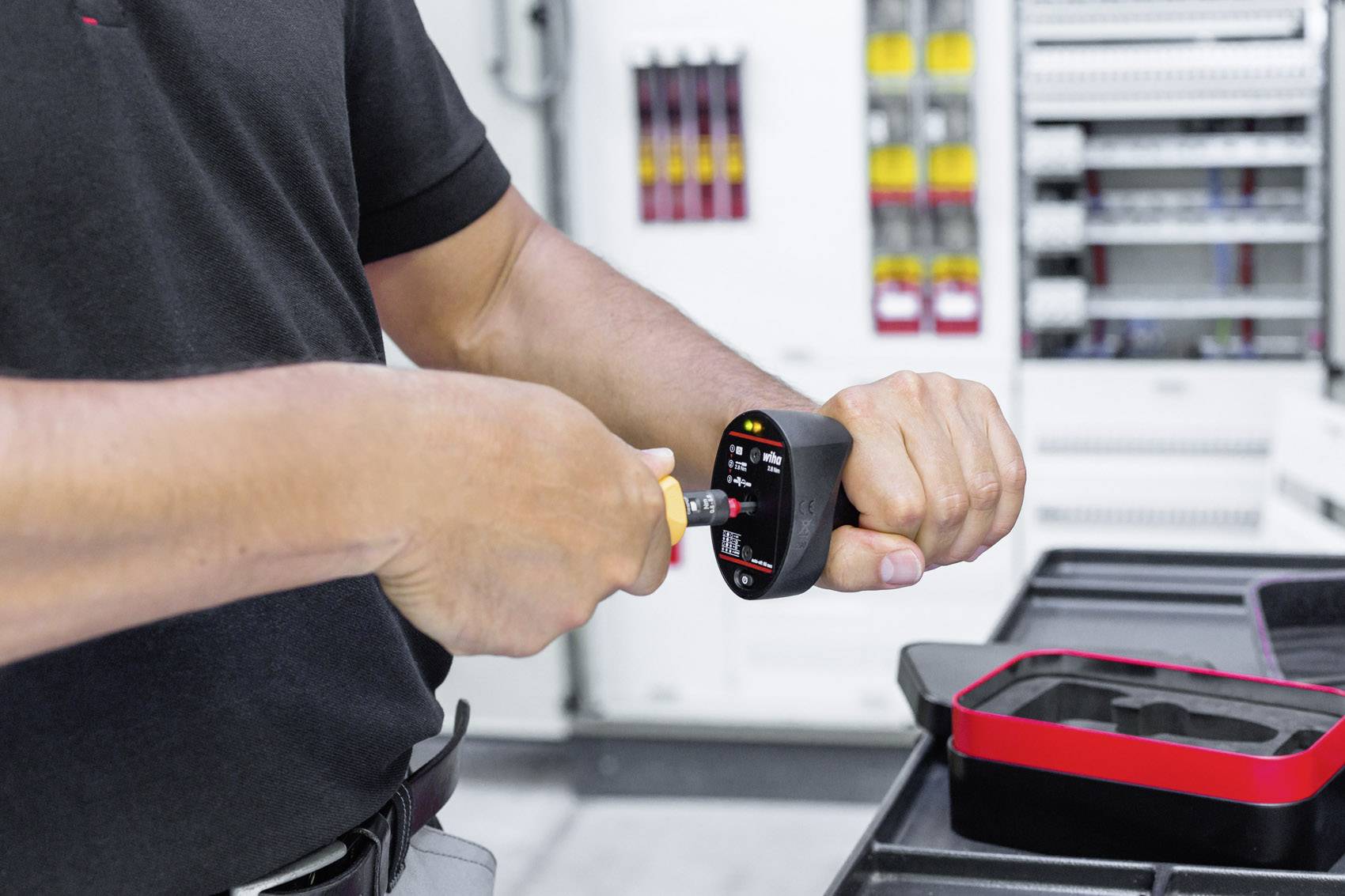 Osoba przeprowadza pomiary za pomocą elektronicznego przyrządu pomiarowego w pomieszczeniu technicznym, w tle widoczne są rozdzielnie elektryczne.