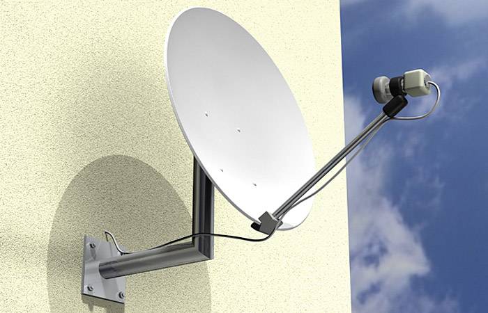 Antena satelitarna jest zamontowana na ścianie budynku i skierowana w niebo. Na tle czystego, błękitnego nieba.