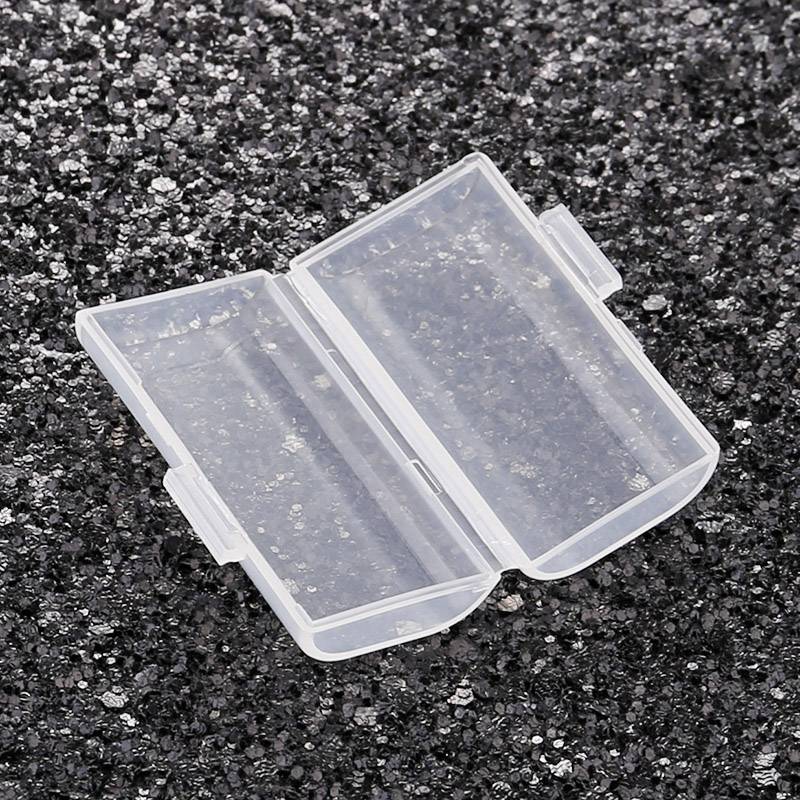 Przezroczysty, prostokątny pojemnik z tworzywa sztucznego leży otwarty na ciemnej, żwirowatej powierzchni.